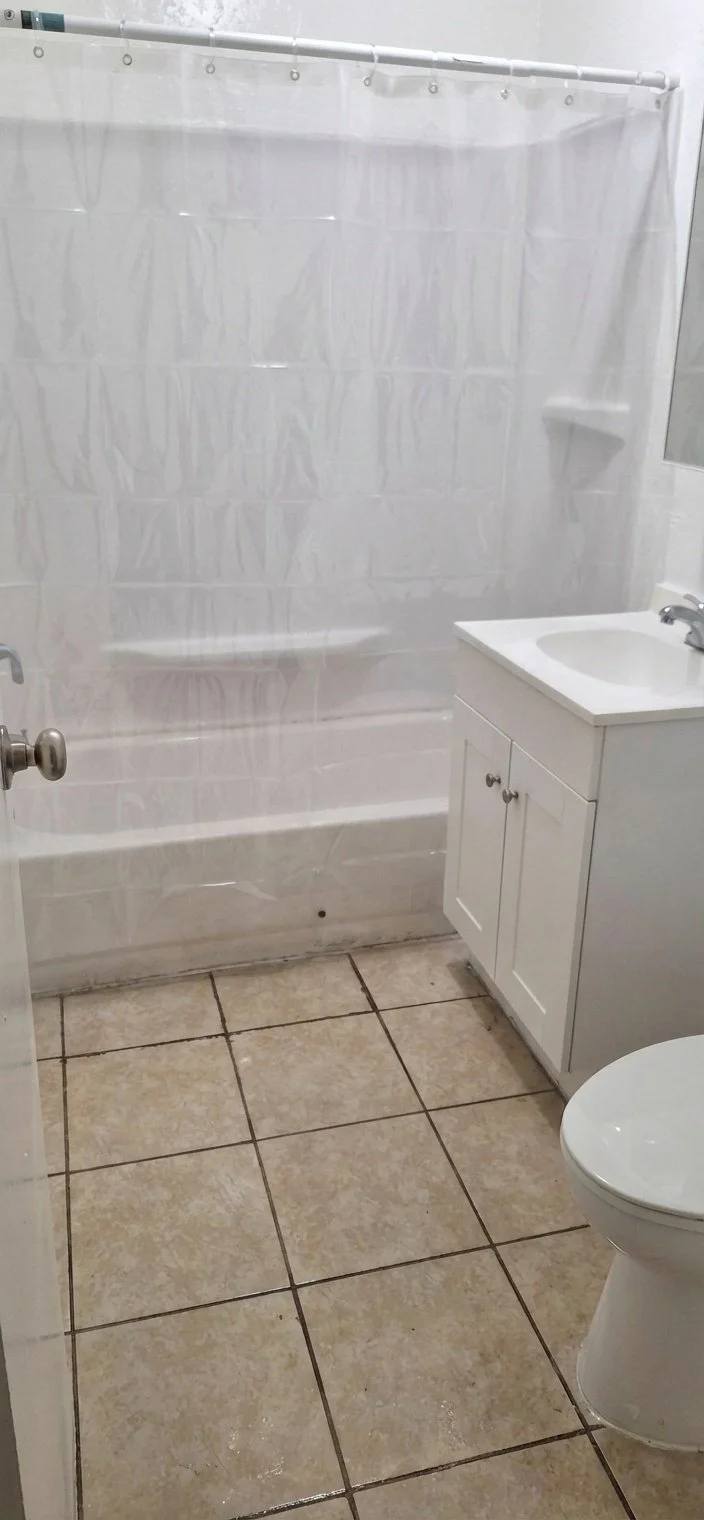 Bathroom with beige tiled floor, white sink vanity, toilet, and shower with a clear plastic curtain.