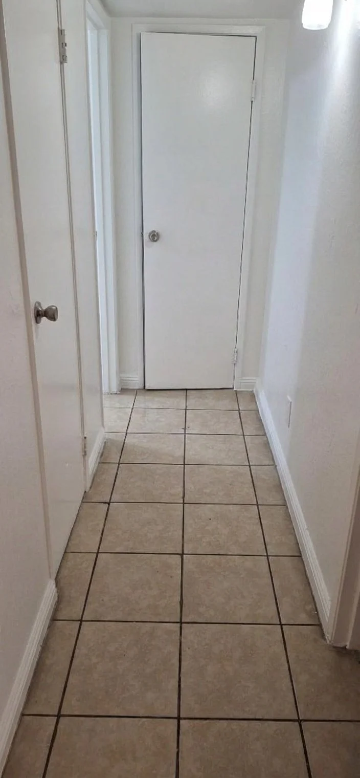 A narrow indoor hallway with beige tiled flooring, white walls, and a closed white door at the end.