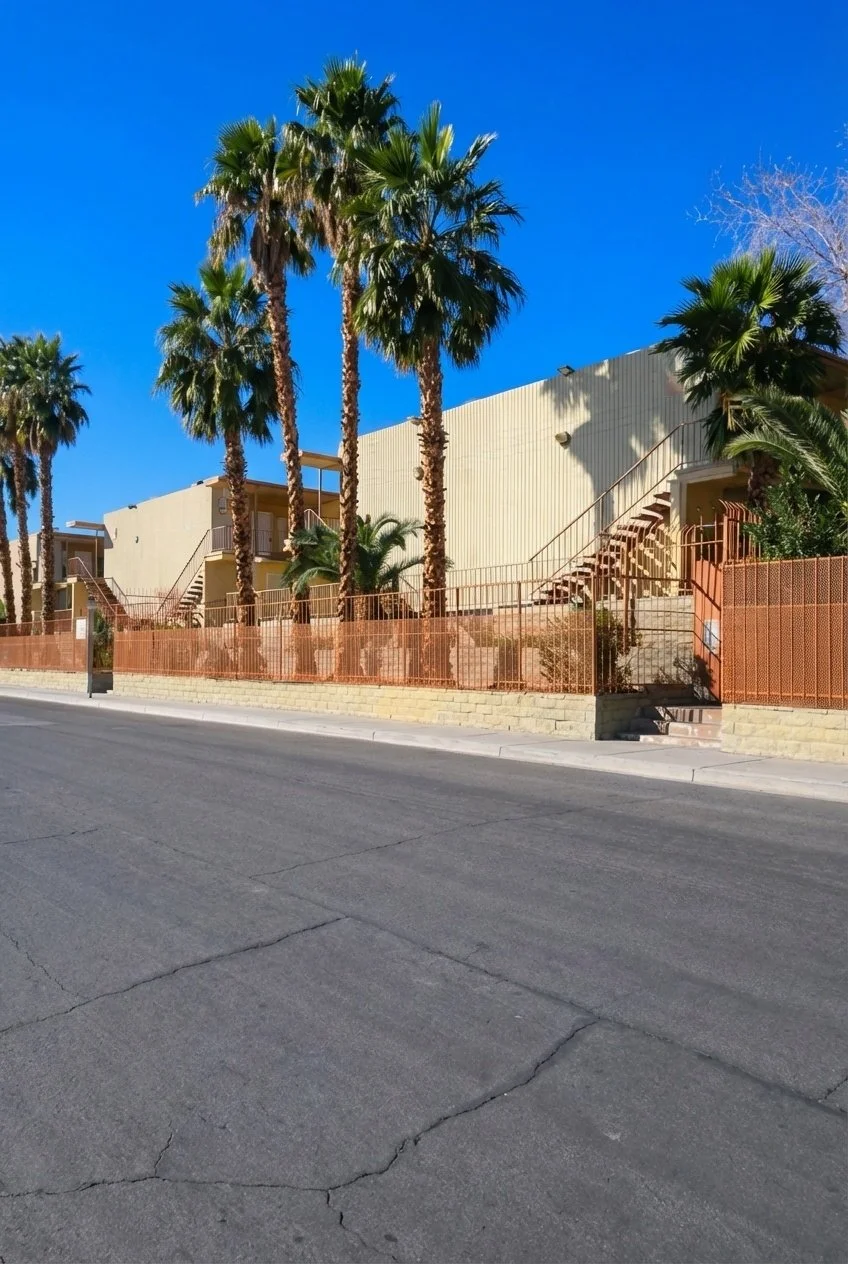 A street view with a building surrounded by tall palm trees, a concrete sidewalk, and a clear blue sky.