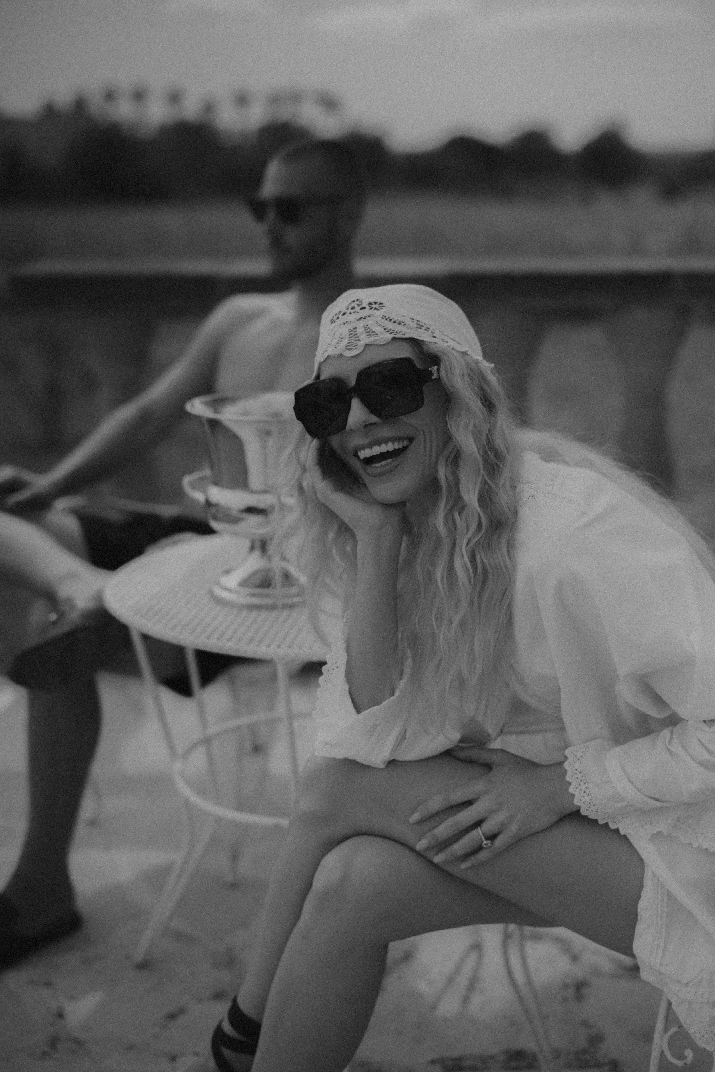Eine lachende Frau mit langen, lockigen Haaren, Sonnenbrille und Sonnenhut im Freien, während sie ein cocktailähnliches Getränk hält. Im Hintergrund sind mehrere Menschen, die vermutlich auf einer Party oder einem sommerlichen Treffen sind.