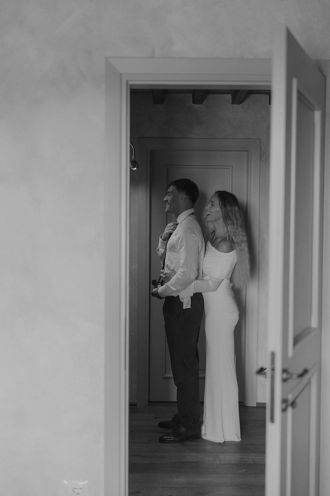 Schwarz-weiß-Foto von einem Paar, das in einem Flur steht, der Mann ist den Damen gestoßen, während sie ihn umarmt, beide lachen und wirken glücklich, im Hintergrund eine Tür.