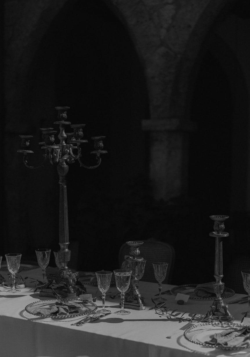 Ein gedeckter Tisch mit Kristallglas, Porzellanschalen, Silberbesteck, dekorativen Kerzenständern und einer Tischdecke, vor einem dunklen, steinernen Hintergrund mit Arkaden.