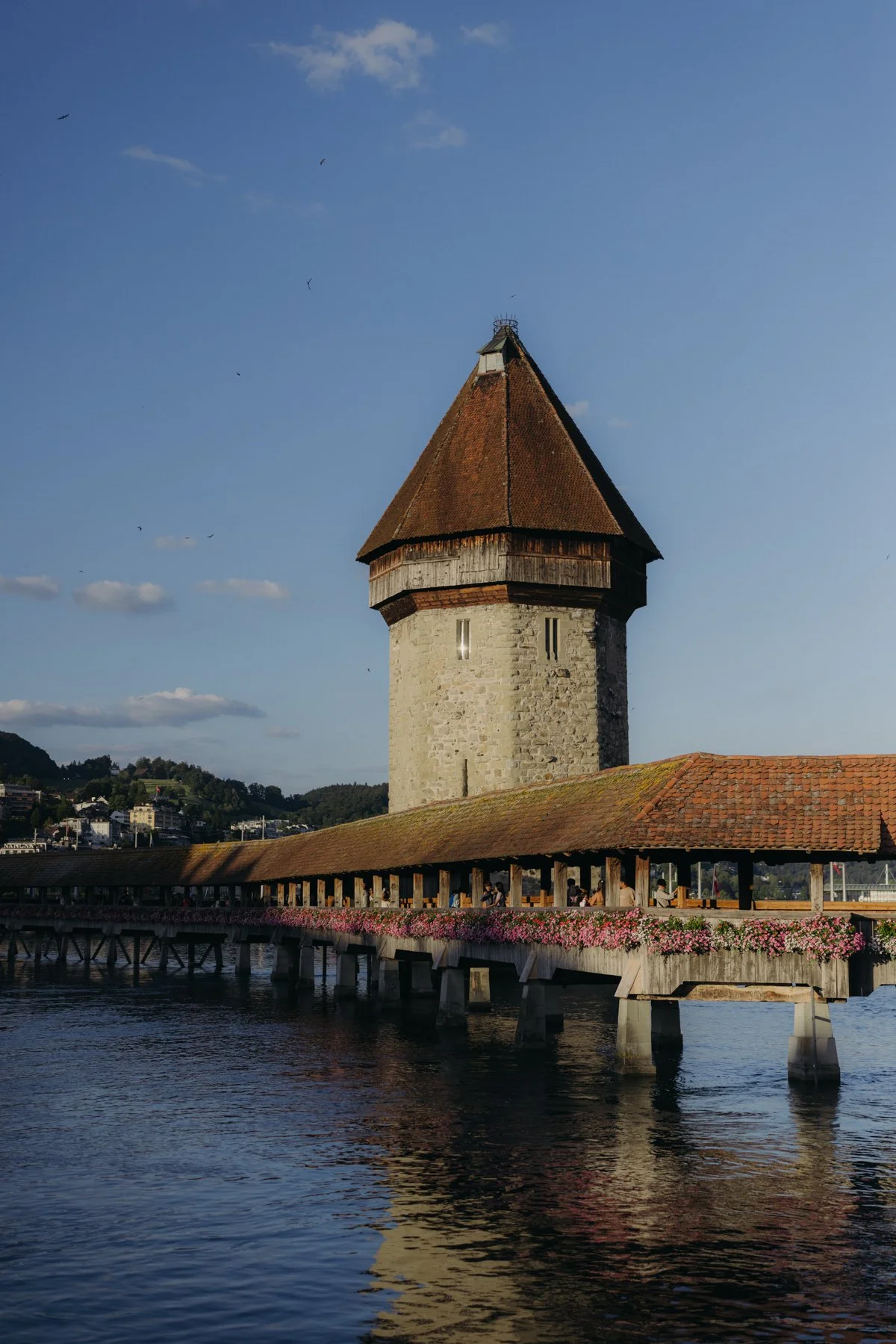 ein alter Wasserturm mit rotem Dach, gelegen an einem Wasserweg mit einer Holzpromenade und Blumen, im Hintergrund eine Stadt und Berge, bewölkter blauer Himmel