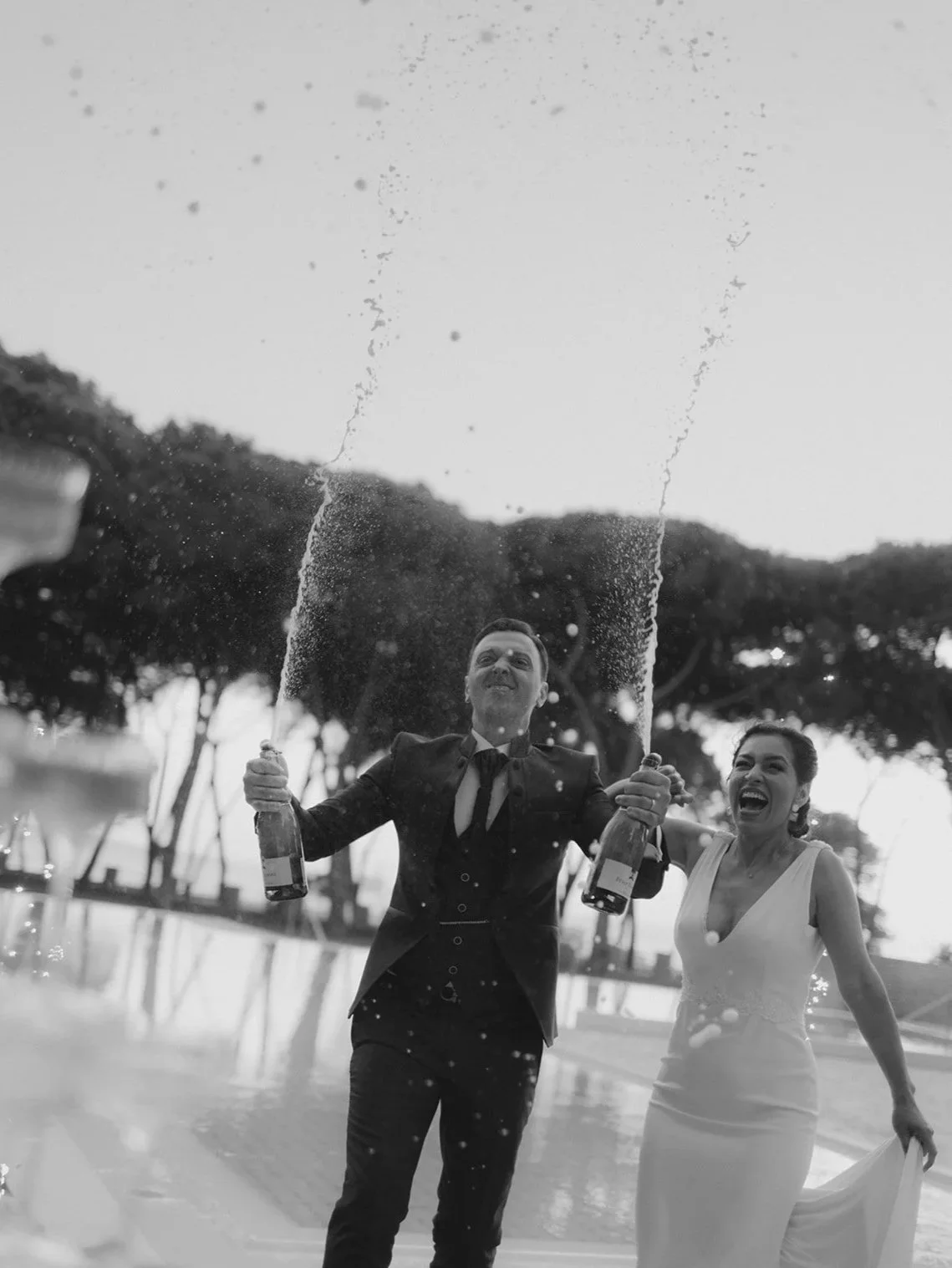 Ein bräutigam in Hochzeitsanzug und eine braut in einem brautKleid feiern mit Sekt, der Wasser spritzt, im Freien, im Schwarz-Weiß-Foto.