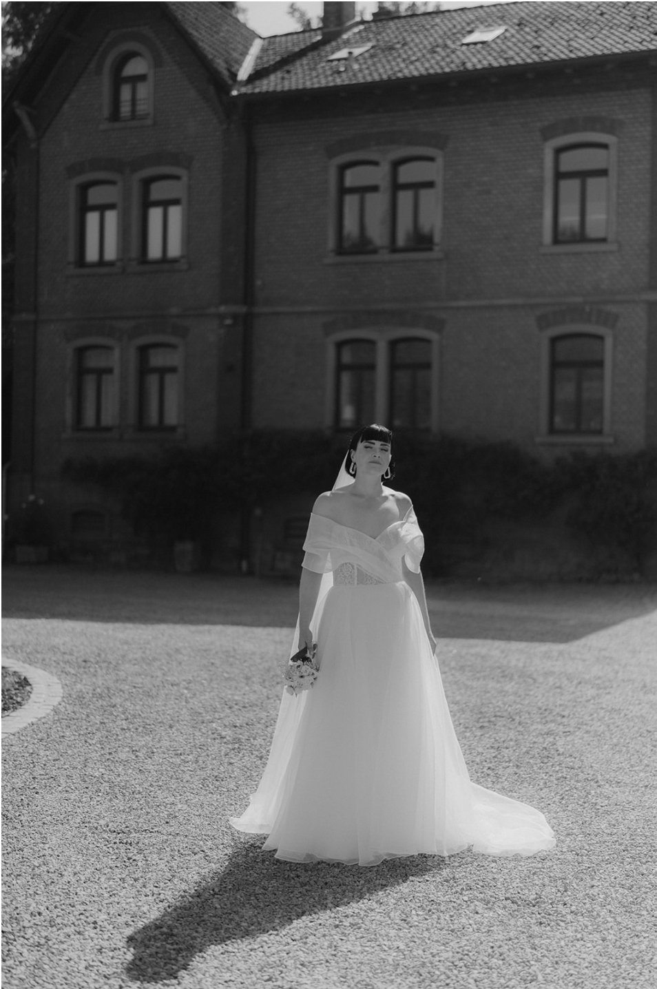 Eine braut in einem weißen Hochzeitskleid steht auf einem Kiesweg vor einem alten Backsteingebäude, das im Sonnenlicht fotografiert wurde.