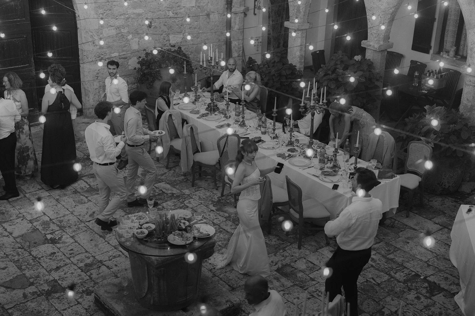 Eine festliche Abendgesellschaft in einem historischen Steingebäude mit Tafel und Kerzen, Menschen, die tanzen, sprechen und trinken bei einer Feier oder Hochzeit.