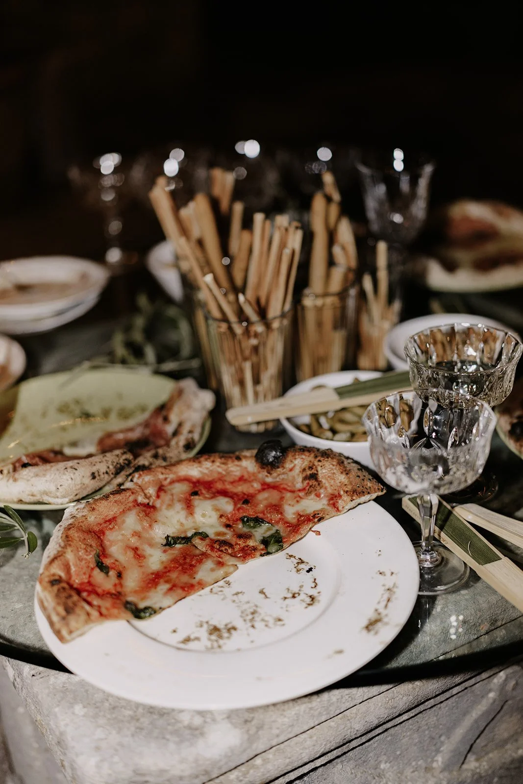 Verwischte Pizza auf einem Teller, Gläser, Brot und mediterrane Speisen auf einem dunklen Tisch bei Abenddämmerung.