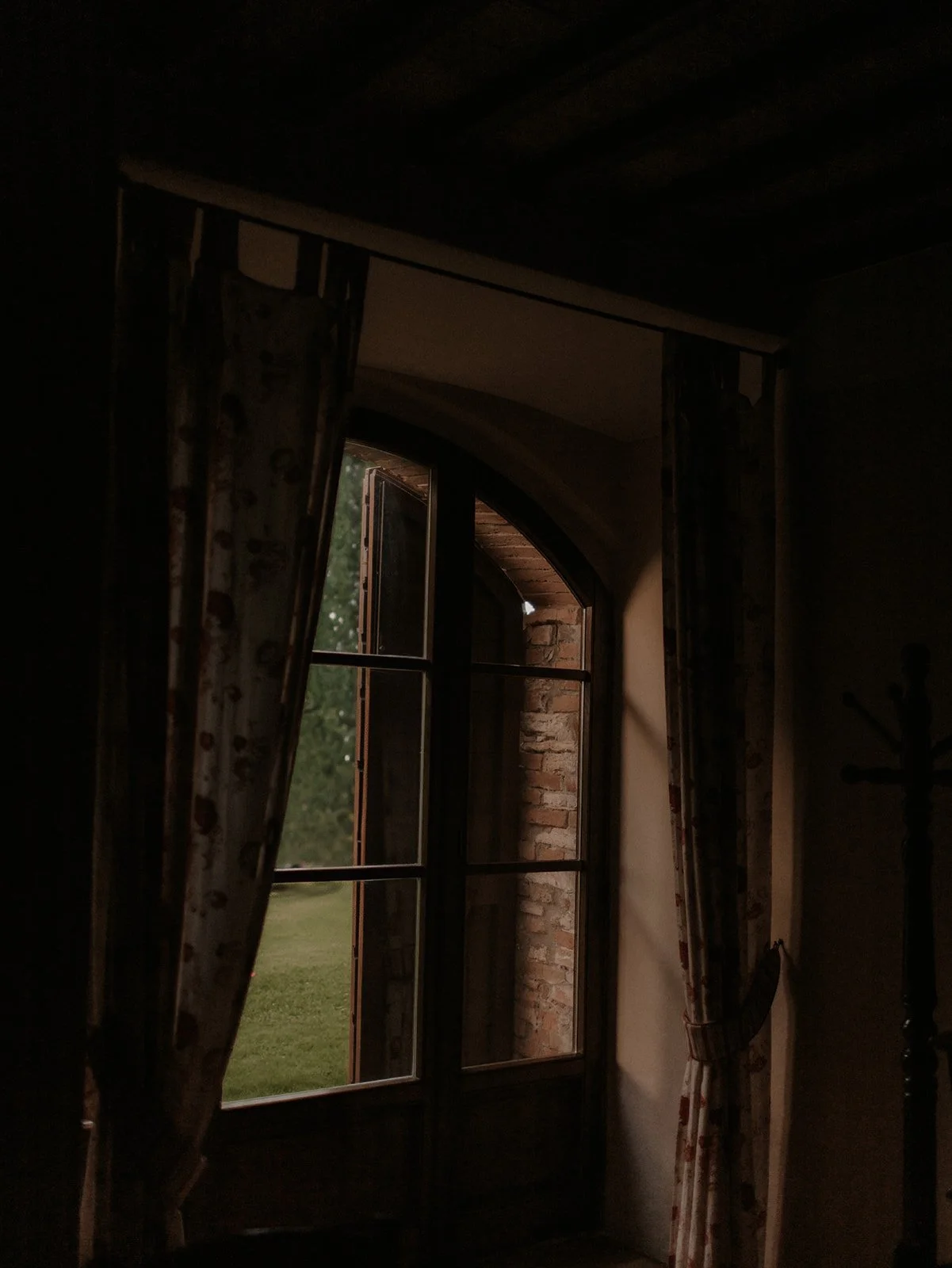 Ein offenes Fenster mit Gardinen in einem dunklen Raum, durch das man einen grünen Garten sieht.