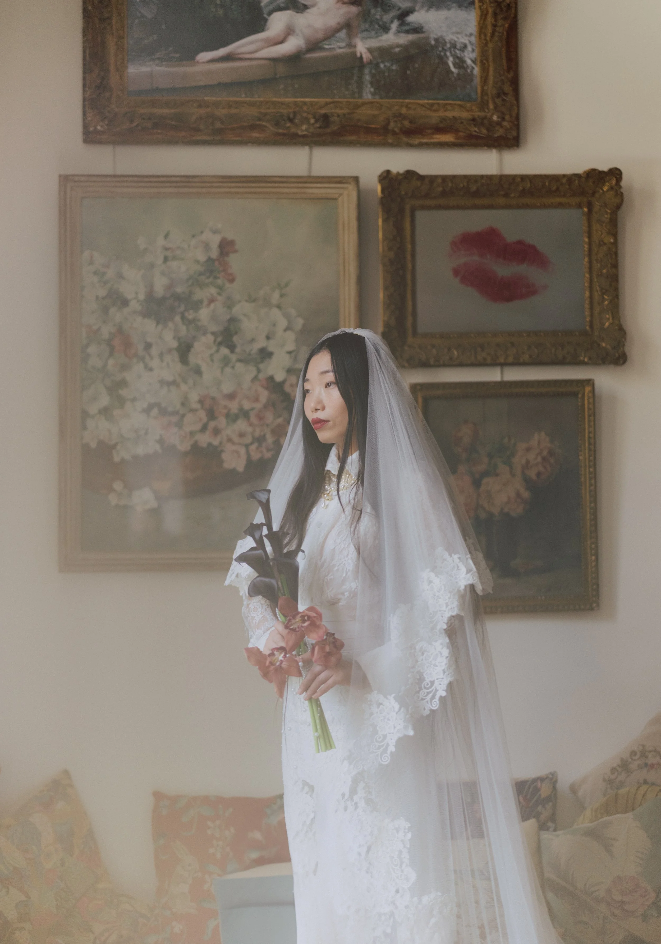 Eine junge Frau in einem weißen Brautkleid mit Schleier hält einen Blumenstrauß und steht in einem Raum mit mehreren Wandbildern, darunter ein Gemälde mit Blumen und eine mit roten Lippen. Sie hat dunkle, lange Haare und einen ruhigen Gesichtsausdruc