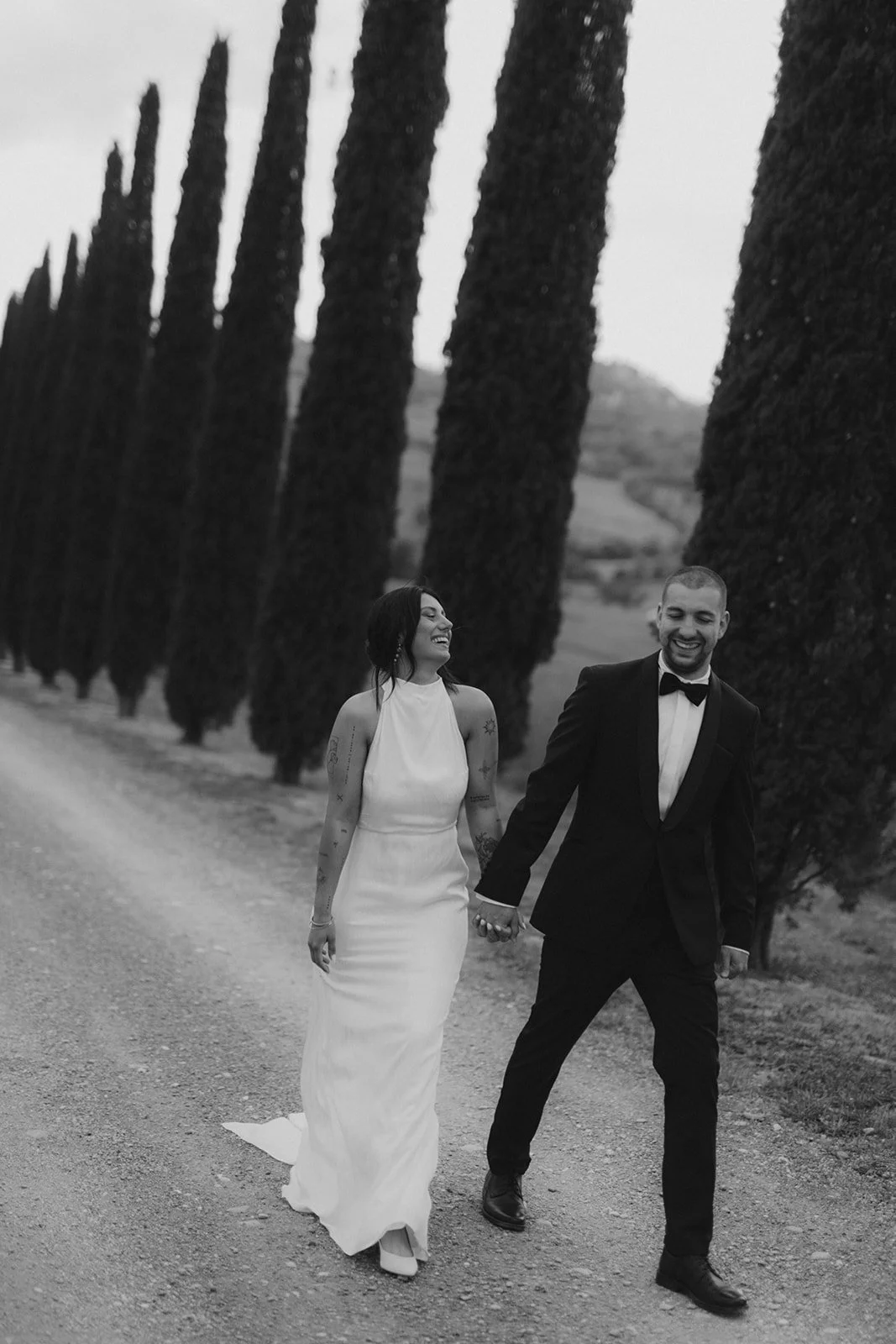 Ein lachendes Paar in formeller Kleidung beim Spaziergang auf einem Kiesweg, umgeben von hohen, schmalen Bäumen, in der Toskana bei Poggio Covili.