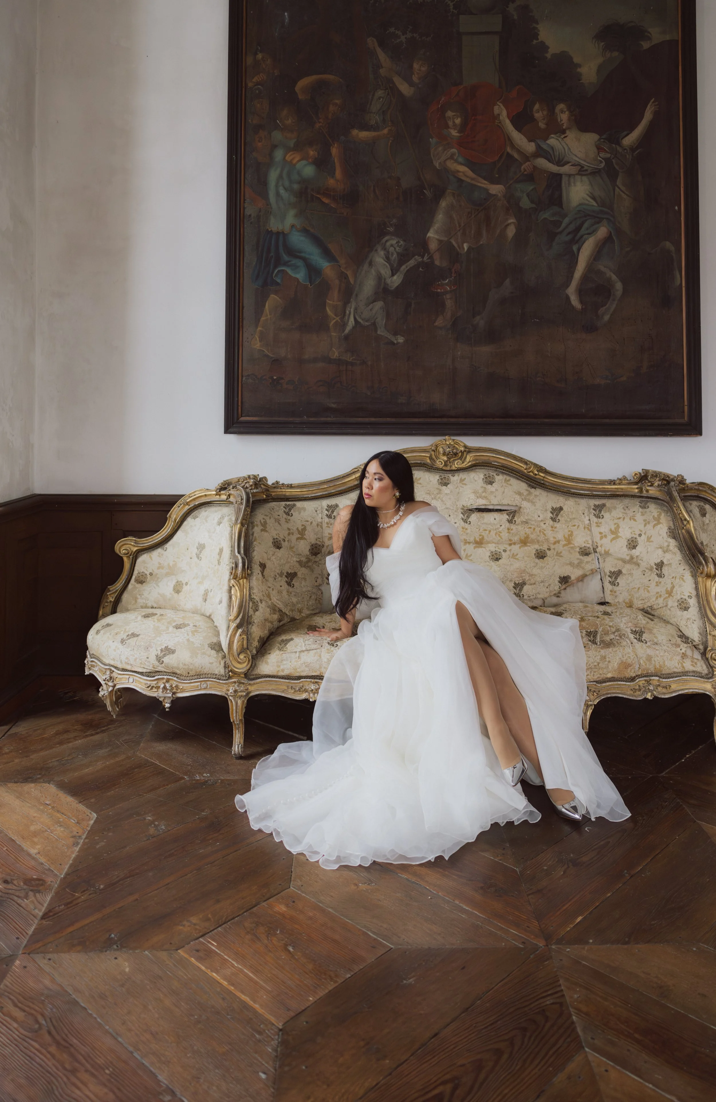 Eine Frau in weißem Brautkleid sitzt auf einer antiken goldfarbenen Couch in einem historischen Raum, im Hintergrund ein großes Gemälde. Editorial bride