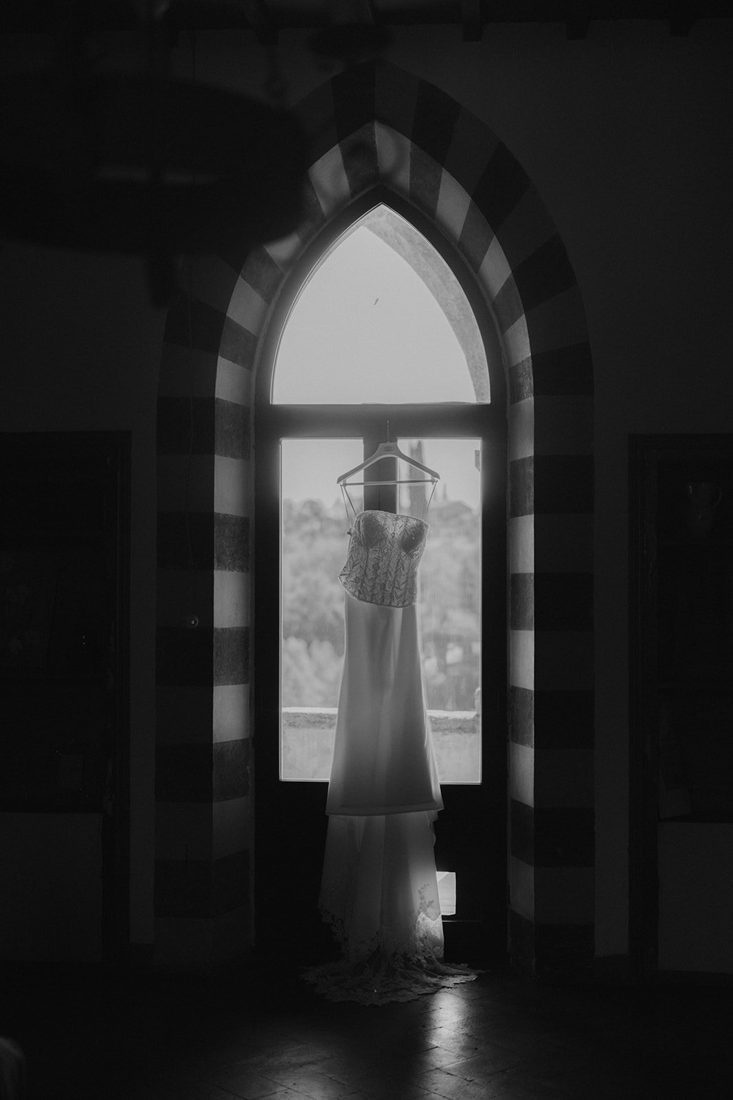 Ein Hochzeitskleid hängt an einem Kleiderbügel vor einem großen, spitzbogigen Fenster, das natürliches Licht hereinlässt.