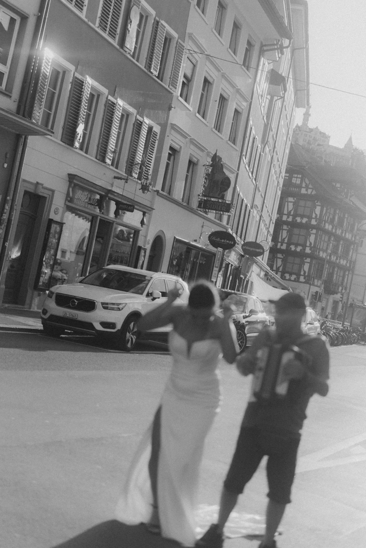 Zwei Frauen stehen auf einer Straße, während im Hintergrund mehrere Autos parken. Die Gebäude sind mehrstöckig und haben viele Fenster, einige mit Fensterläden. Es sind auch Geschäfte in der Straße sichtbar.