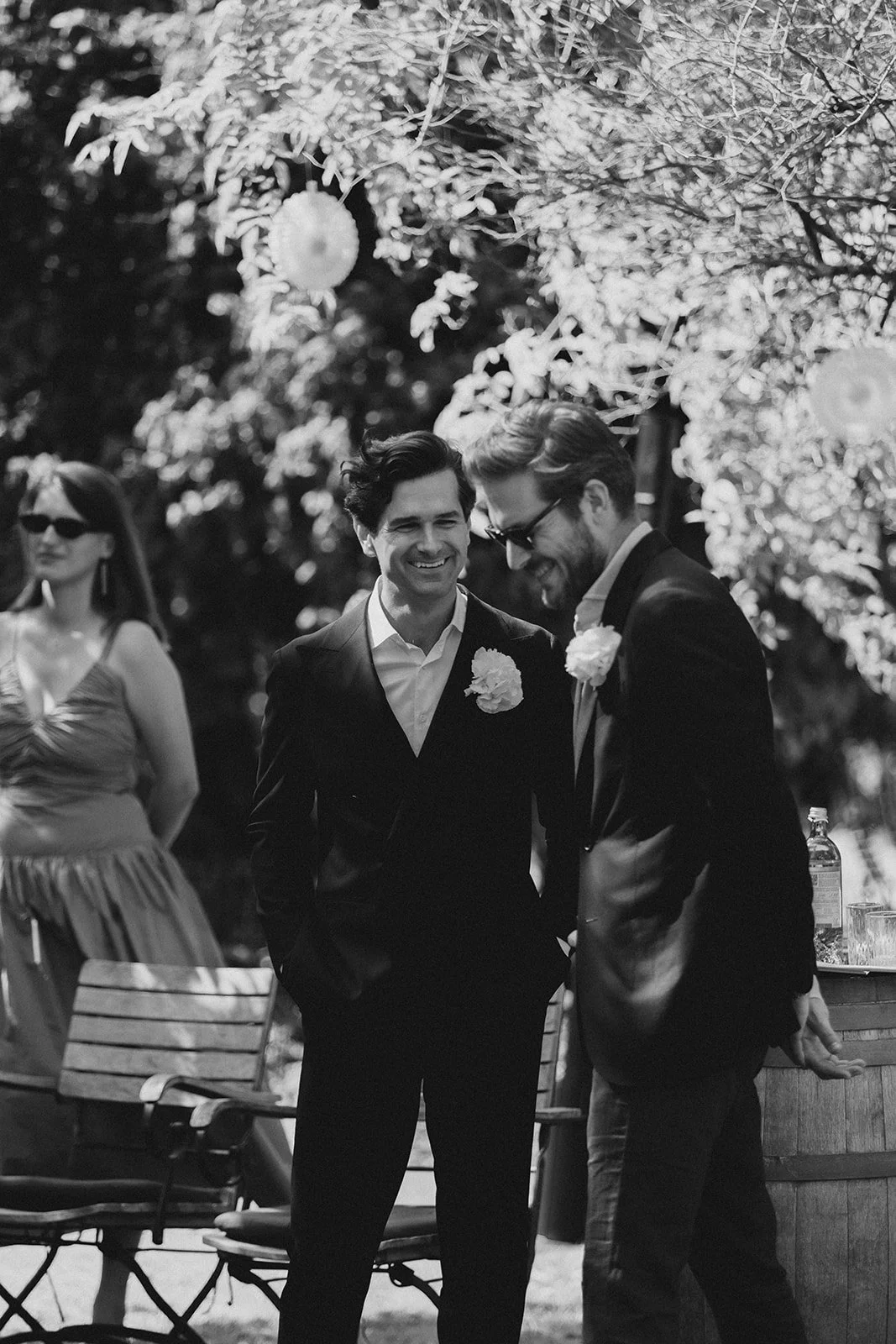 Zwei Männer in Anzügen mit Blumen im Revers, die lächelnd und im Gespräch bei einer Outdoor-Hochzeit stehen, im Hintergrund eine Frau und dekorative Lampions. Bräutigam und Trauzeuge In der Schamarimühle