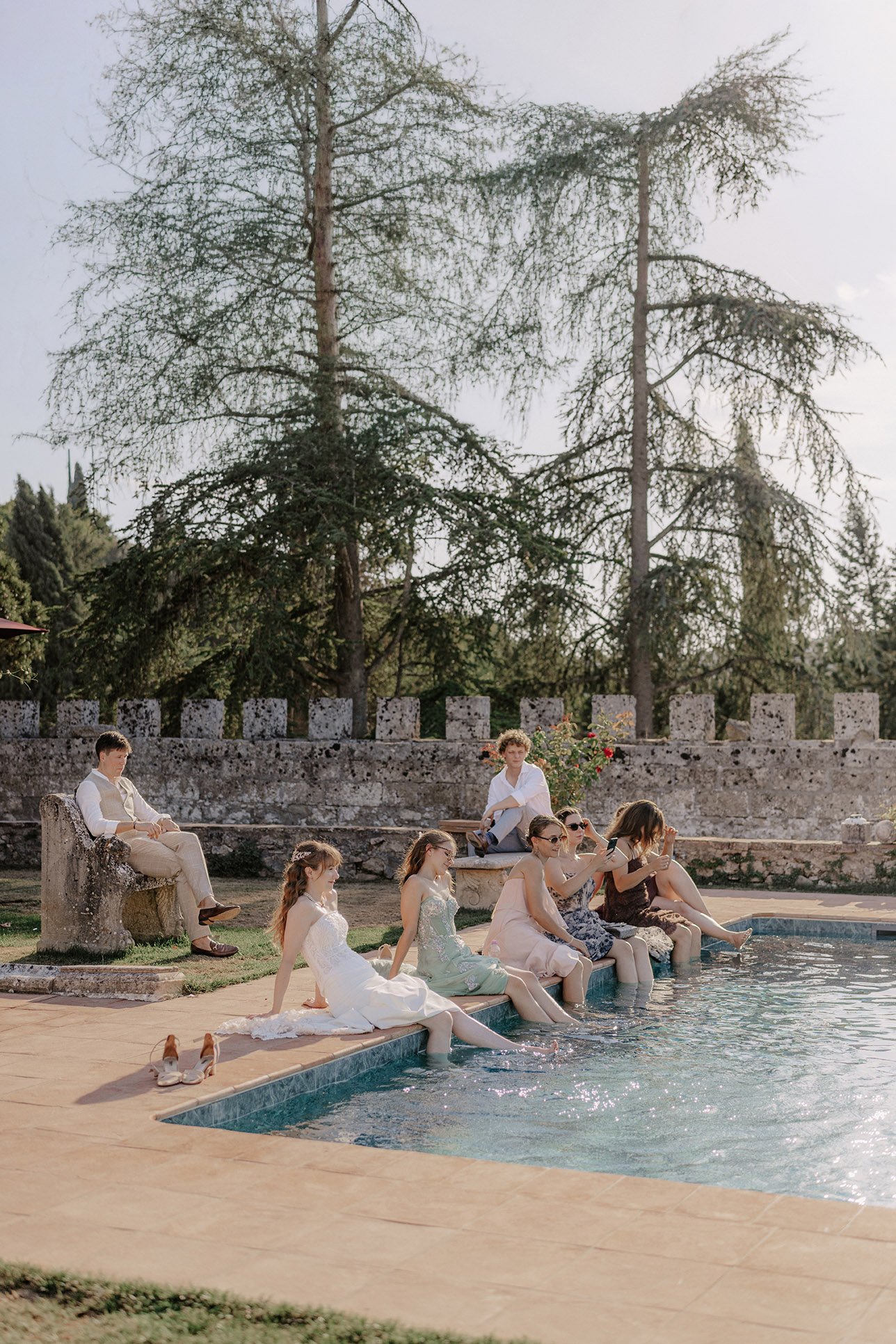 Eine Gruppe von Menschen, die an einem Pool sitzen, einige im Wasser und andere auf der Poolkante, bei Sonnenuntergang in einem Garten mit großen Bäumen im Hintergrund. Es scheint eine festliche oder besondere Gelegenheit zu sein.