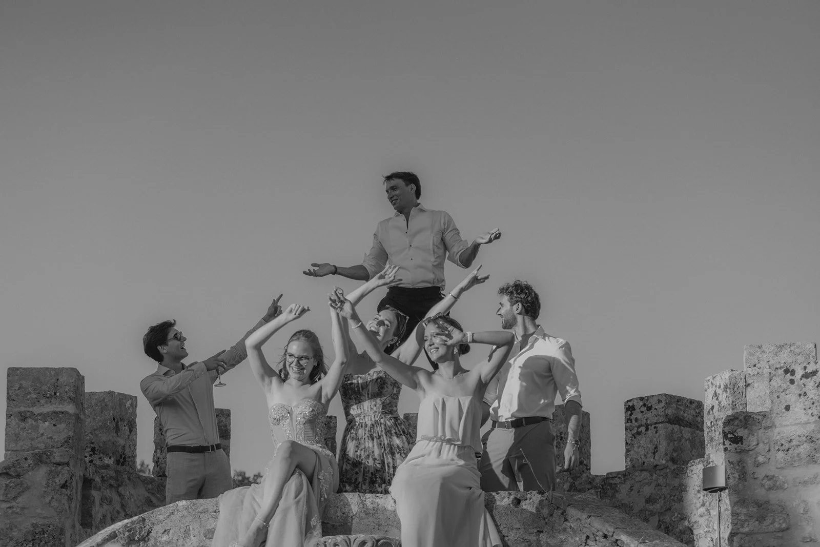 Eine Gruppe von sechs Menschen, davon drei sitzend und drei stehend, lachen und feiern auf einer alten Steinmauer im Freien bei Sonnenuntergang. Ein Mann sitzt auf den Schultern eines anderen Mannes.