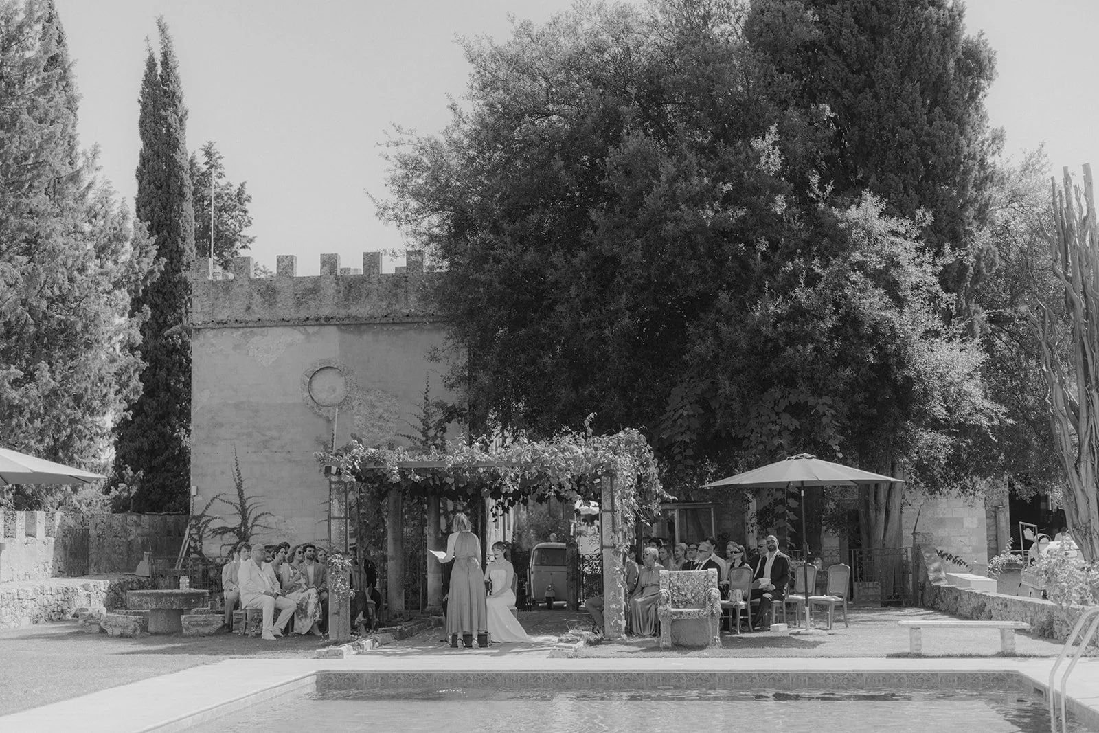 Eine Hochzeit im Freien auf einer italienischen Villa vor einer alten Steinmauer, mit Gästen, eleganten Stühlen, einem Pavillon, einer Poolseite und großen Bäumen im Hintergrund.