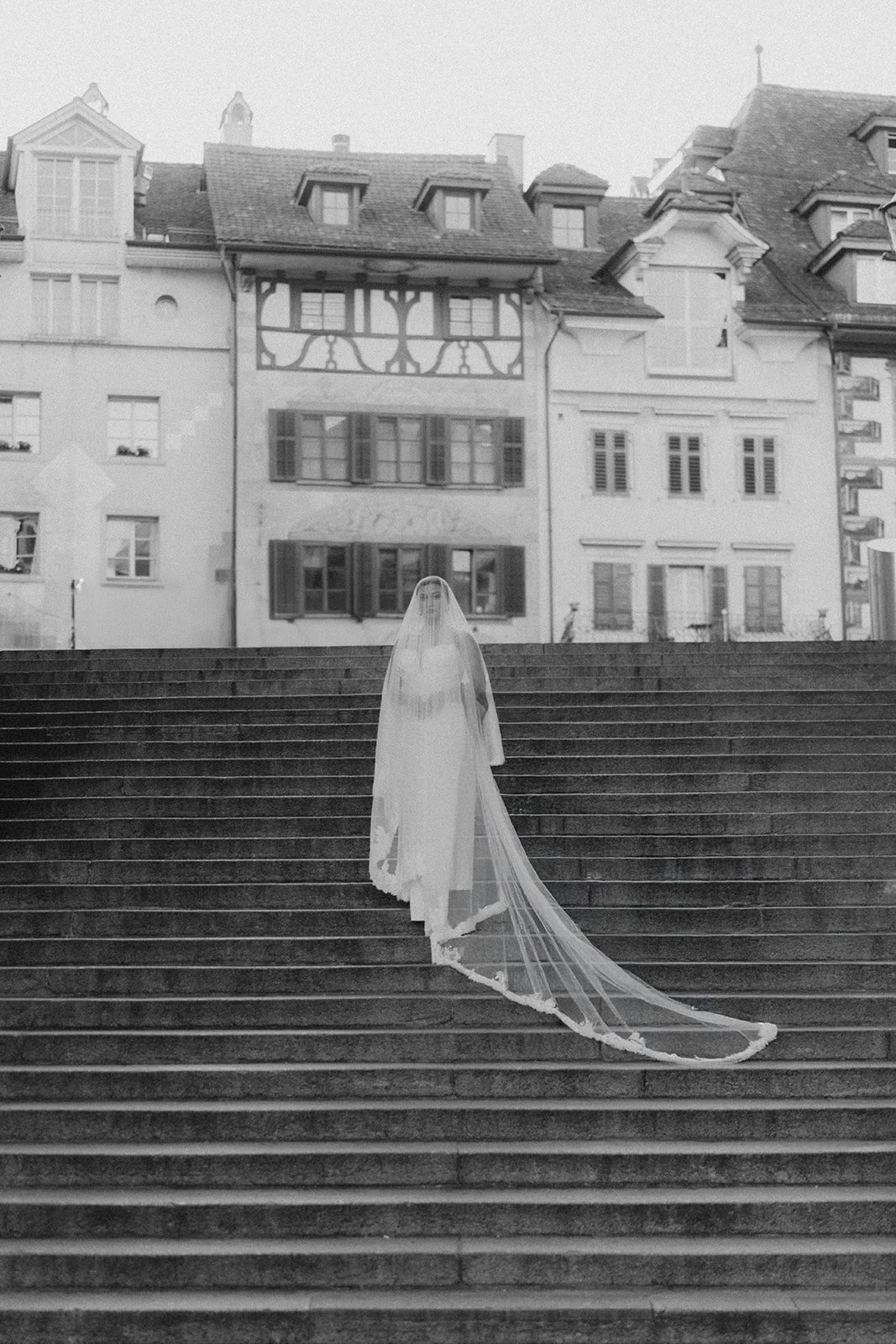 Braut in einem Hochzeitskleid mit Schleier auf Treppe vor historischen Gebäuden.