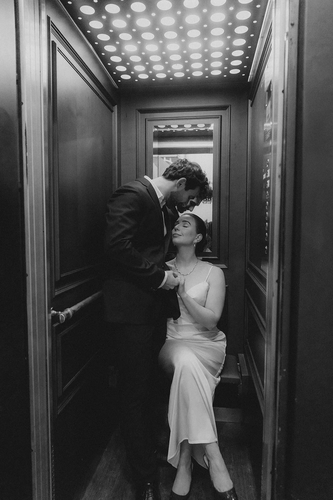 Ein Bräutigam und eine Braut in HochzeitsKleidung, die in einem Aufzug posieren. Der Mann trägt einen dunklen Anzug und die Frau ein langes, helles Kleid. Sie sehen sich liebevoll an, während die Frau auf einem Sitz im Aufzug sitzt.
