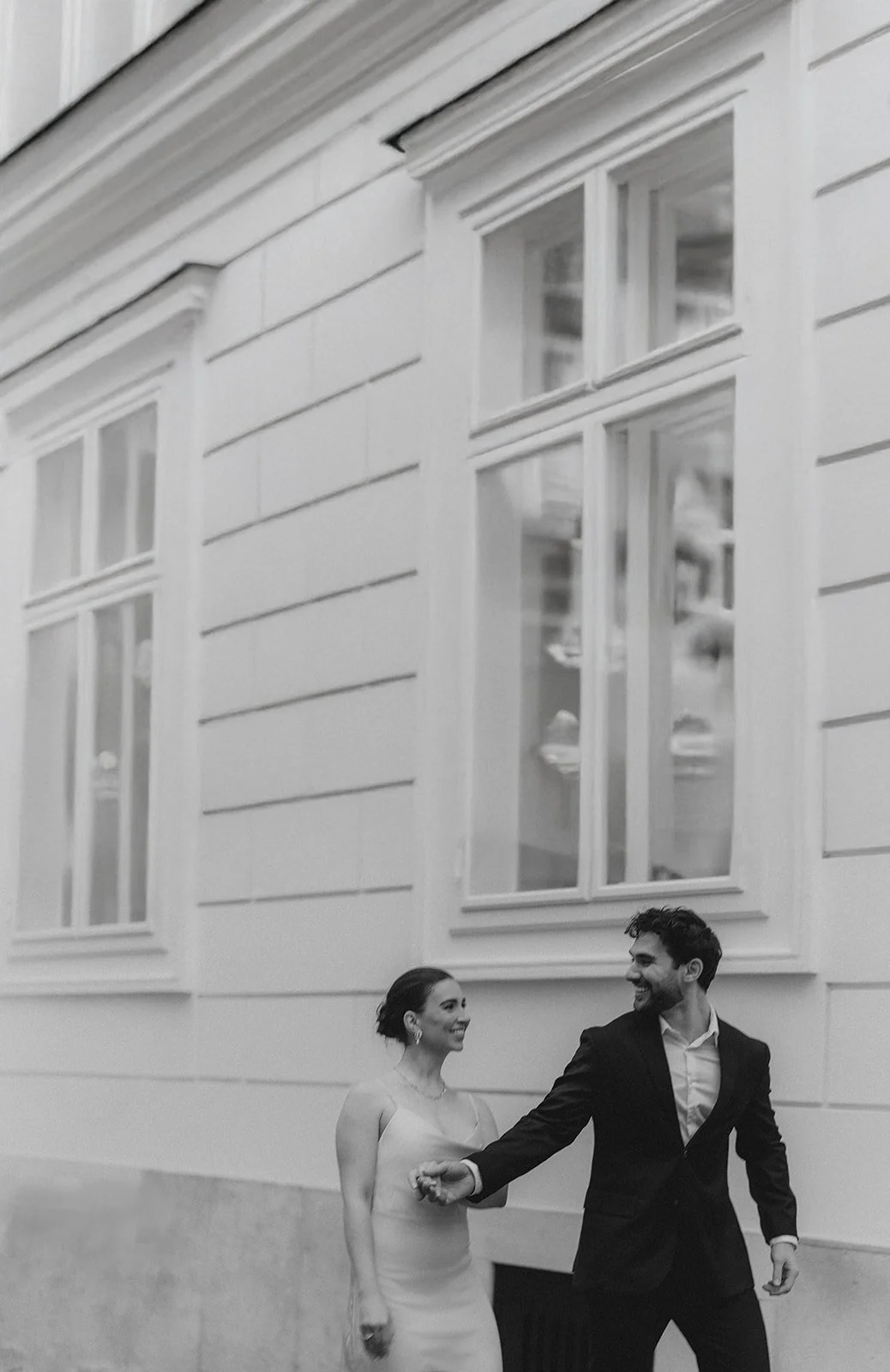 Ein lachendes Paar, ein Mann in Anzug und eine Frau in Kleid, tanzen vor einem weißen Gebäude mit großen Fenstern in Wiener Altstadt. Bridalcouple