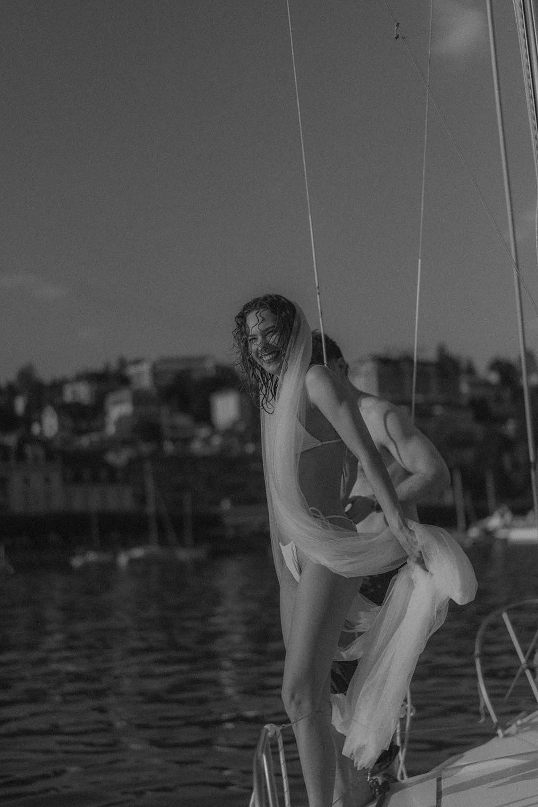 Frau mit nassem Haar, lachend, auf einem Boot im Wasser, trägt ein Tuch. Im Hintergrund eine Stadtansicht am Ufer.