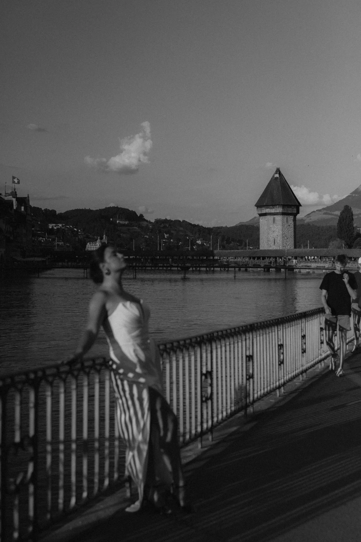 Schwarz-weiß-Foto von einer Frau in einem weißen Top und gestreiften Hosen, die an einem Flussufer steht, mit einem historischen Turm im Hintergrund und einem Mann, der auf einem Gehweg steht.