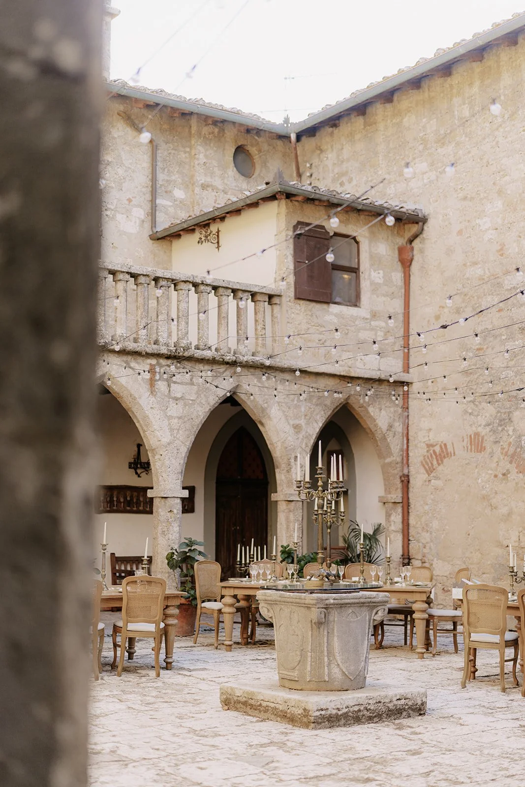 Ein Innenhof mit einem Steintisch und Stühlen, Dekorationsstücken wie Kerzenleuchter und Pflanzen, umgeben von einer alten Steinmauer und einem mehrstöckigen Gebäude mit Fensterläden und einer Balustrade, festliche Lichterkette über dem Bereich.