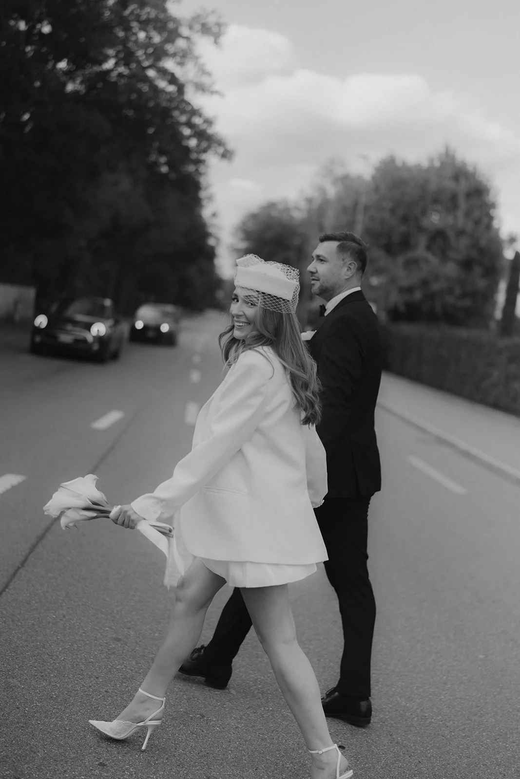 Ein Paar im Hochzeitsoutfit steht in der Mitte einer Straße, mit dem Rücken zum Betrachter, während die Frau lachend in Richtung Kamera schaut und die Straße entlang läuft, ohne Schuhe, hält einen Blumenstrauß in der Hand, im Hintergrund sind Autos u