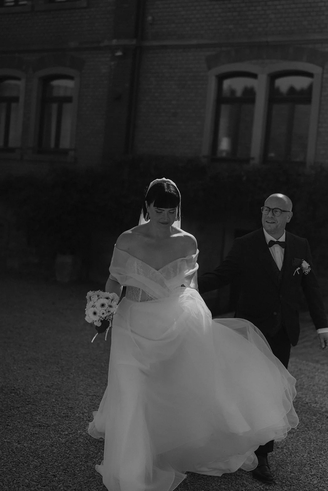Schwarz-Weiß-Foto eines Brautpaares bei ihrer Hochzeit, die Braut trägt ein off-the-Shoulder-Kleid und hält einen Blumenstrauß, der Bräutigam trägt einen Anzug mit Fliege, sie gehen Hand in Hand auf dem Gutshof unterbessenbach