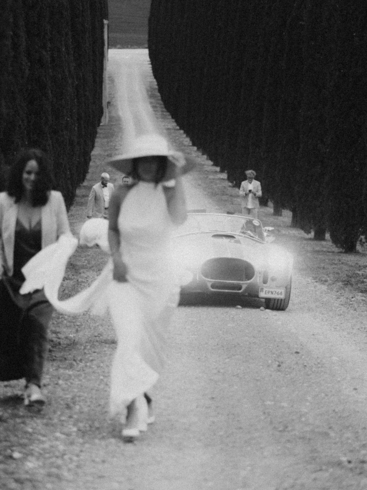 Menschen in eleganter Kleidung gehen auf einer Landstraße, im Hintergrund ein Sportwagen. Es ist eine Schwarz-Weiß-Fotografie mit einer Reihe hoher, gepflegter Bäume auf beiden Seiten der Straße.