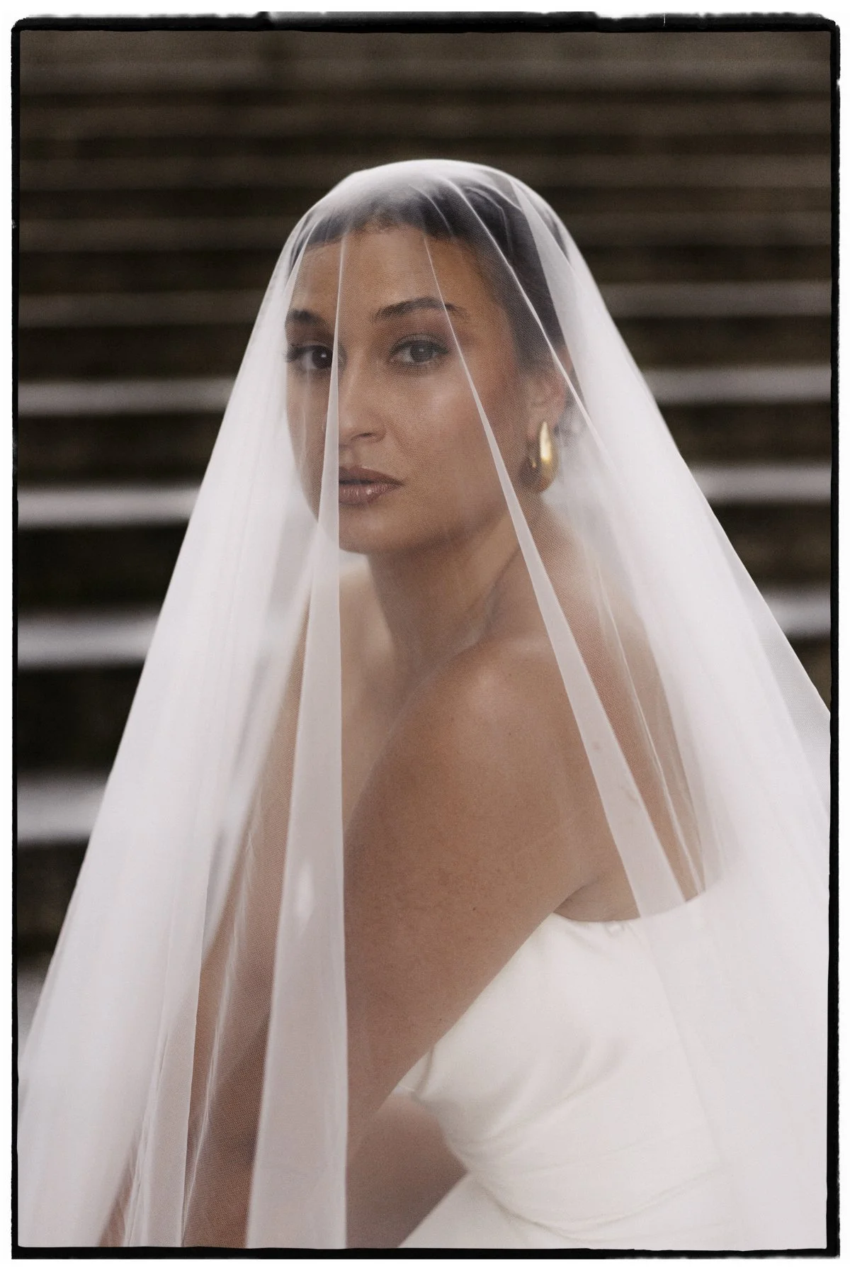 Frau mit Brautkleid und Schleier, blauer Augen und goldenen Ohrringen, auf steinigen Stufen sitzend.