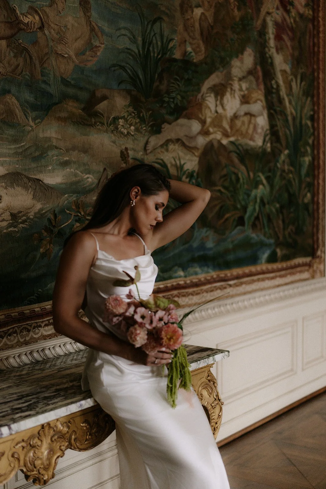 Eine Braut in einem weißen Kleid hält einen Blumenstrauß und steht vor einer goldverzierten Wand und einem Wandteppich in einem opulenten Raum des St charles Hall in Meggen in der Schweiz an ihrer hochzeit
