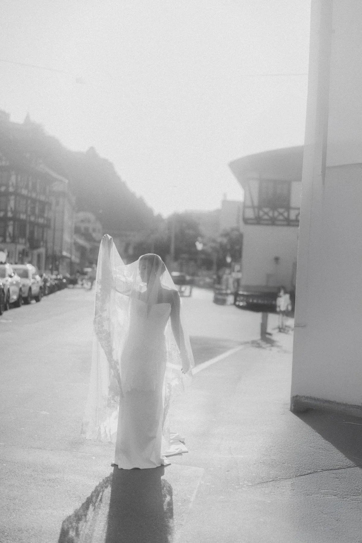 Eine Frau in einem weißen Brautkleid mit Schleier steht auf einer Straße in der Sonne, im Hintergrund sind Gebäude und Fahrzeuge sichtbar.