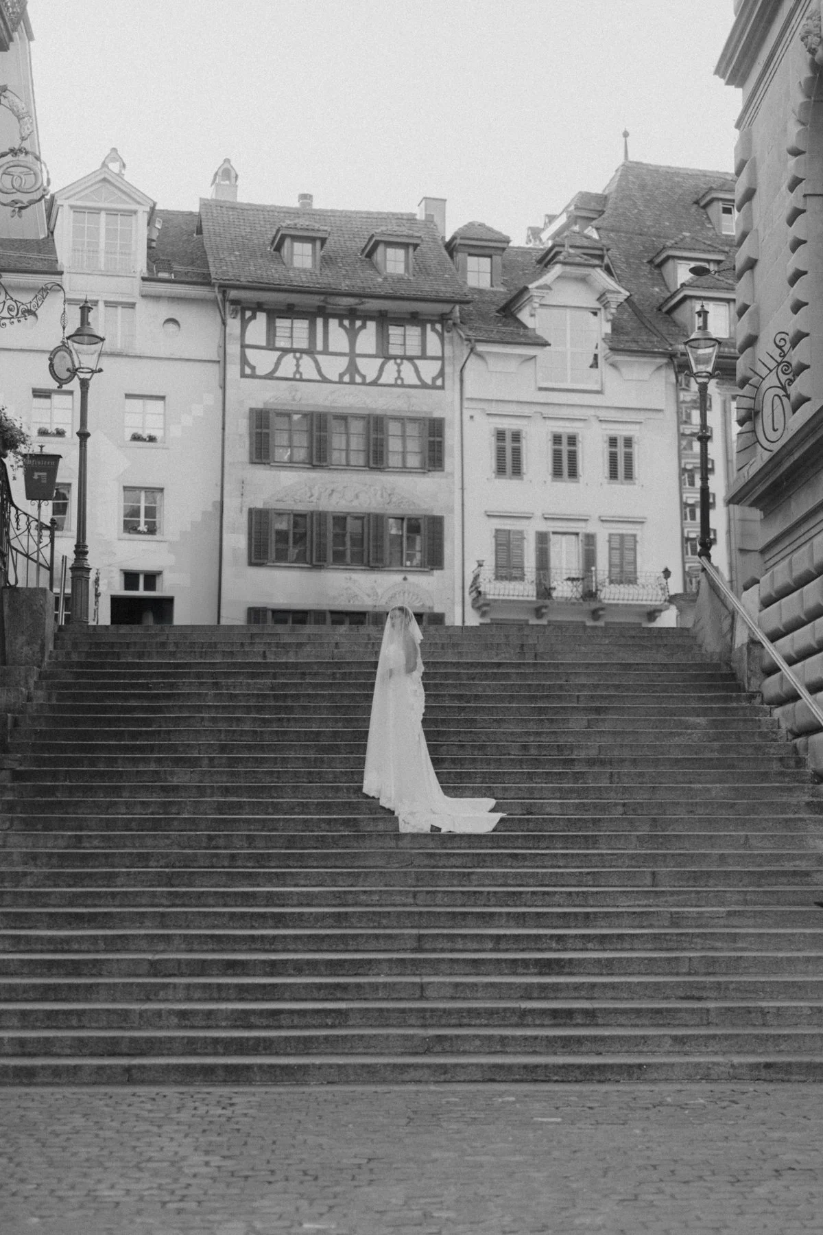 Schwarz-weiß-Foto einer Frau in einem Brautkleid, die auf einer Treppe steht, umgeben von historischen Gebäuden mit klassischen Lampen, vermutlich bei einer Hochzeitsfotografie.