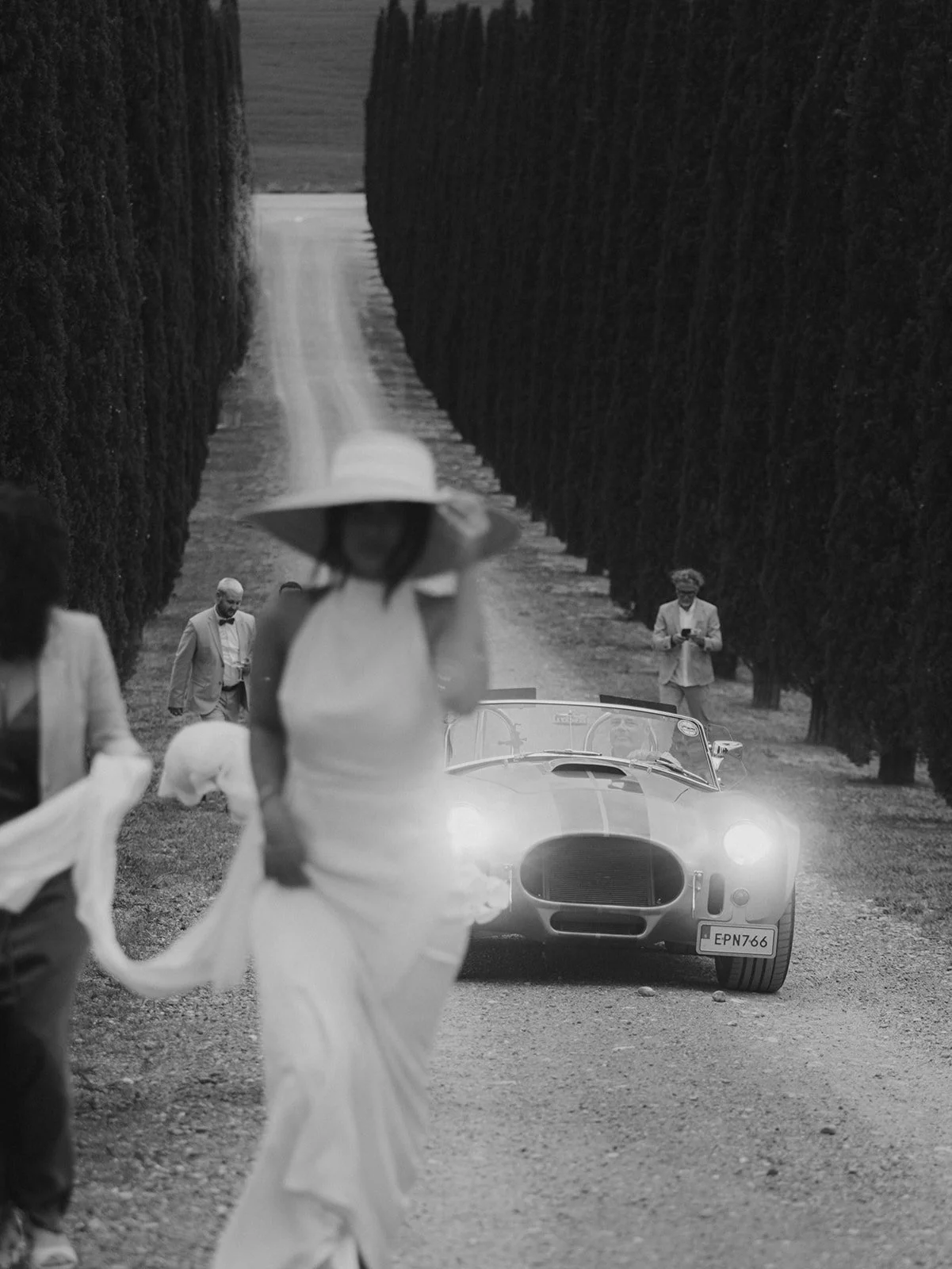 Schwarz-Weiß-Fotografie einer Hochzeitsfeier im Freien, im Vordergrund eine Braut in einem Brautkleid, im Hintergrund ein klassisches Auto, in der Auffahrt von Poggio Covili