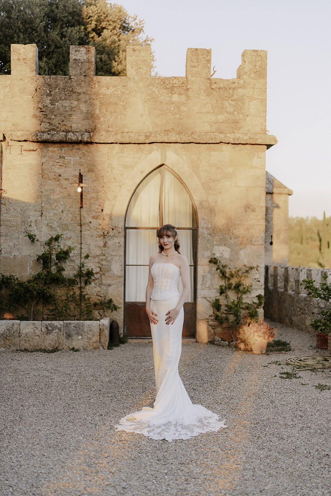 Eine Frau in einem weißen Hochzeitskleid steht vor einer alten Steingebäude mit einem großen Fenster. Es ist sunset, und die Szene ist ruhig und malerisch.