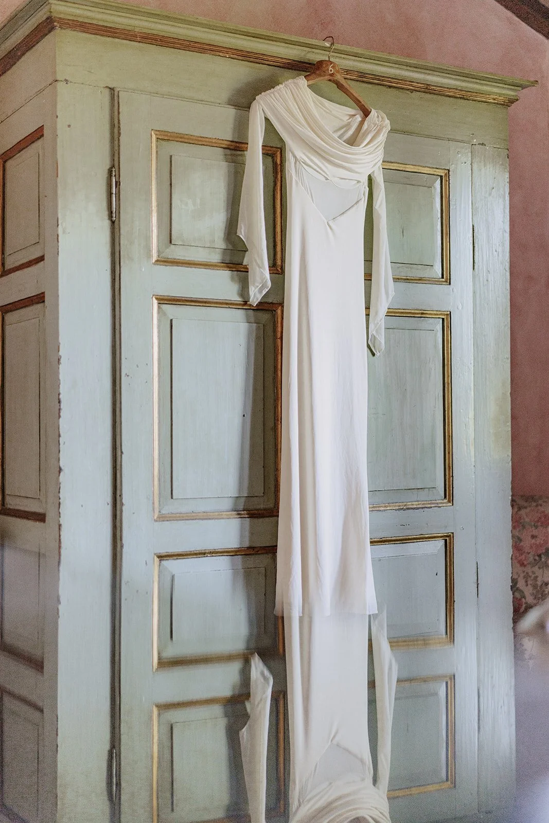 Weiße Brautkleid hängt an einem Holzkleiderbügel an einer alten grünen Holztür, im Hintergrund eine pinkfarbene Wand.