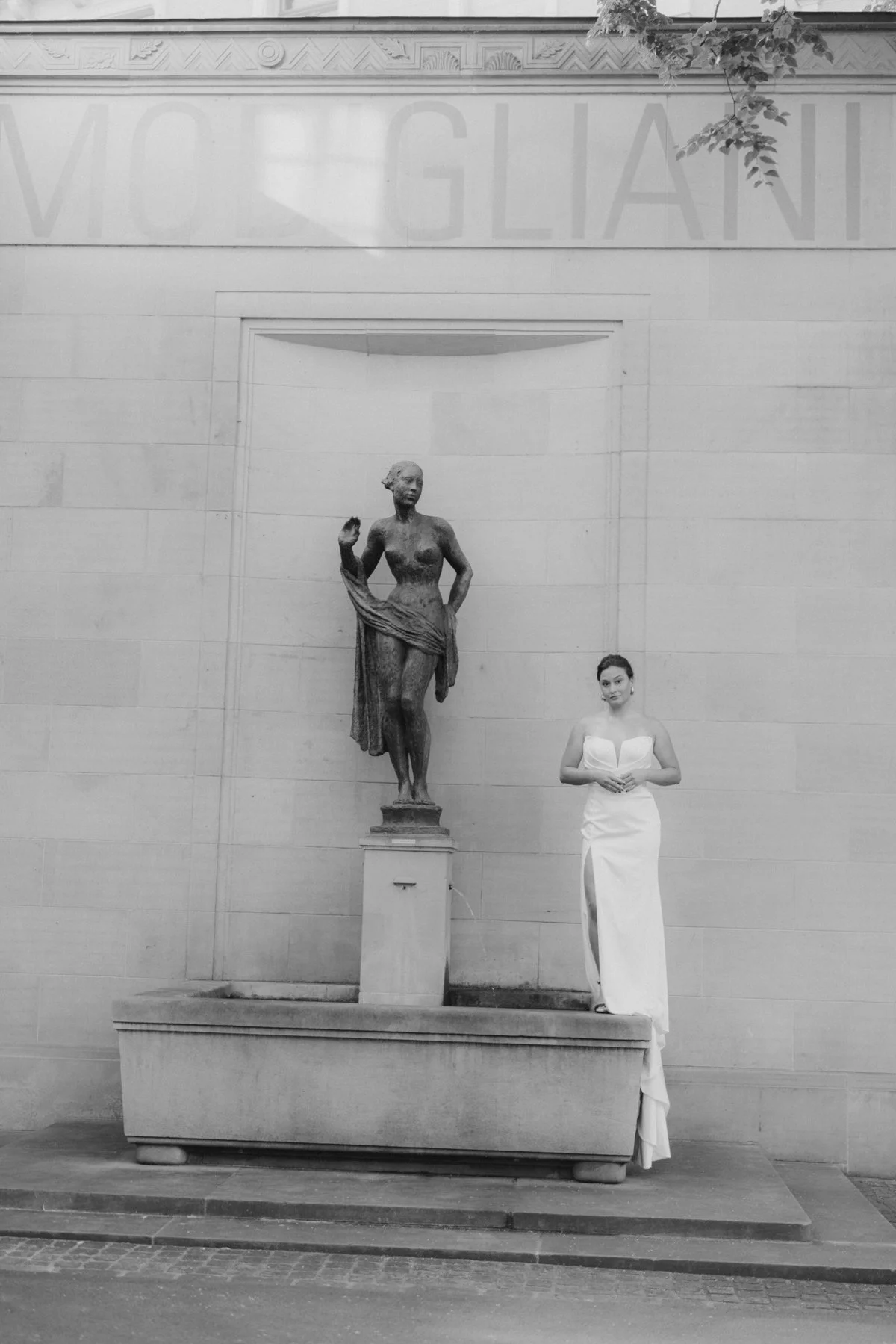 Schwarz-Weiß-Foto einer Statue einer nackten Frau, die auf einem Sockel steht, und einer Frau in einem eleganten weißen Abendkleid neben der Statue. Im Hintergrund eine Wand mit einer Inschrift. Über der Inschrift einige Blätter einer Pflanze.