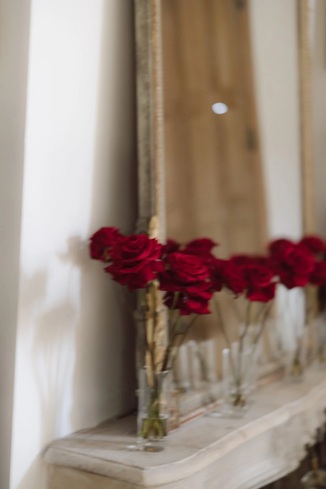 Rote Rosen in kleinen Glaskaraffen vor einem großen Spiegel, der auf einem cremefarbenen Tisch steht.