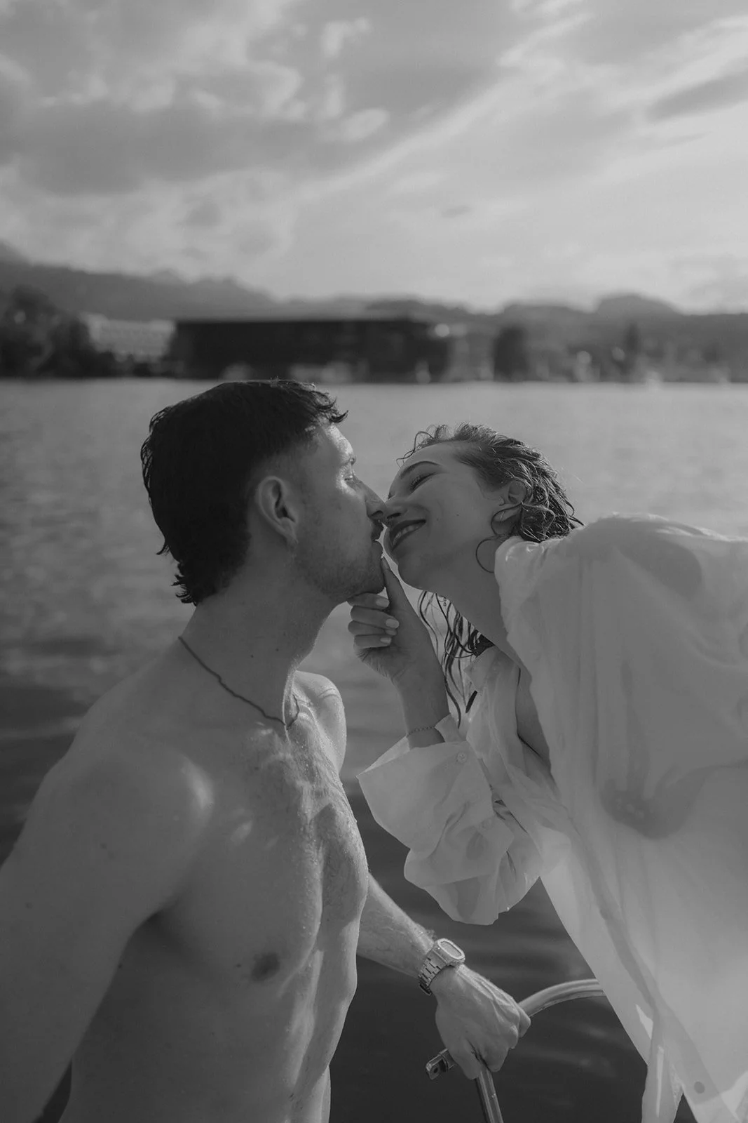 Ein Mann und eine Frau, die sich nahe am Wasser beim Schmusen befinden, amüsiert sich, während sie sich küssen, und lachen in einer entspannten, schönen Szenerie am See oder Fluss bei Sonnenuntergang, in Schwarz-Weiß.