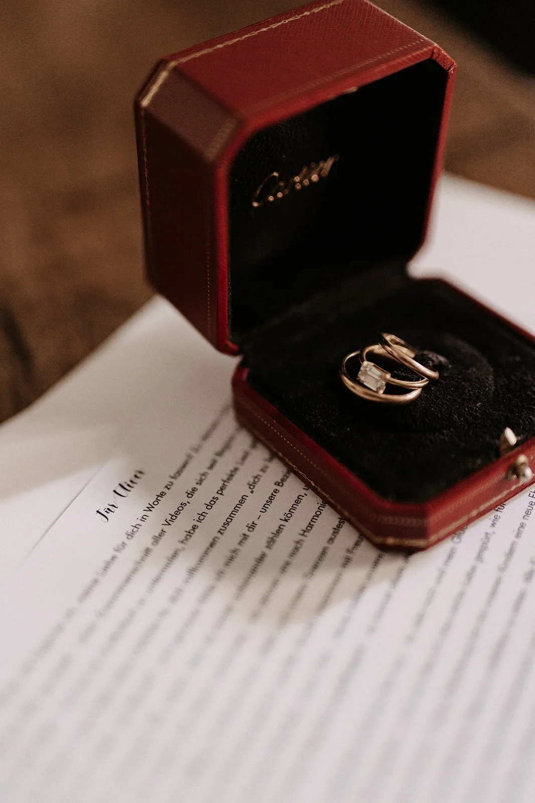 Rote Schmuckschatulle mit zwei Ringen und einem kleinen Edelstein innen, auf einem weißen Papier mit Text in Deutsch.