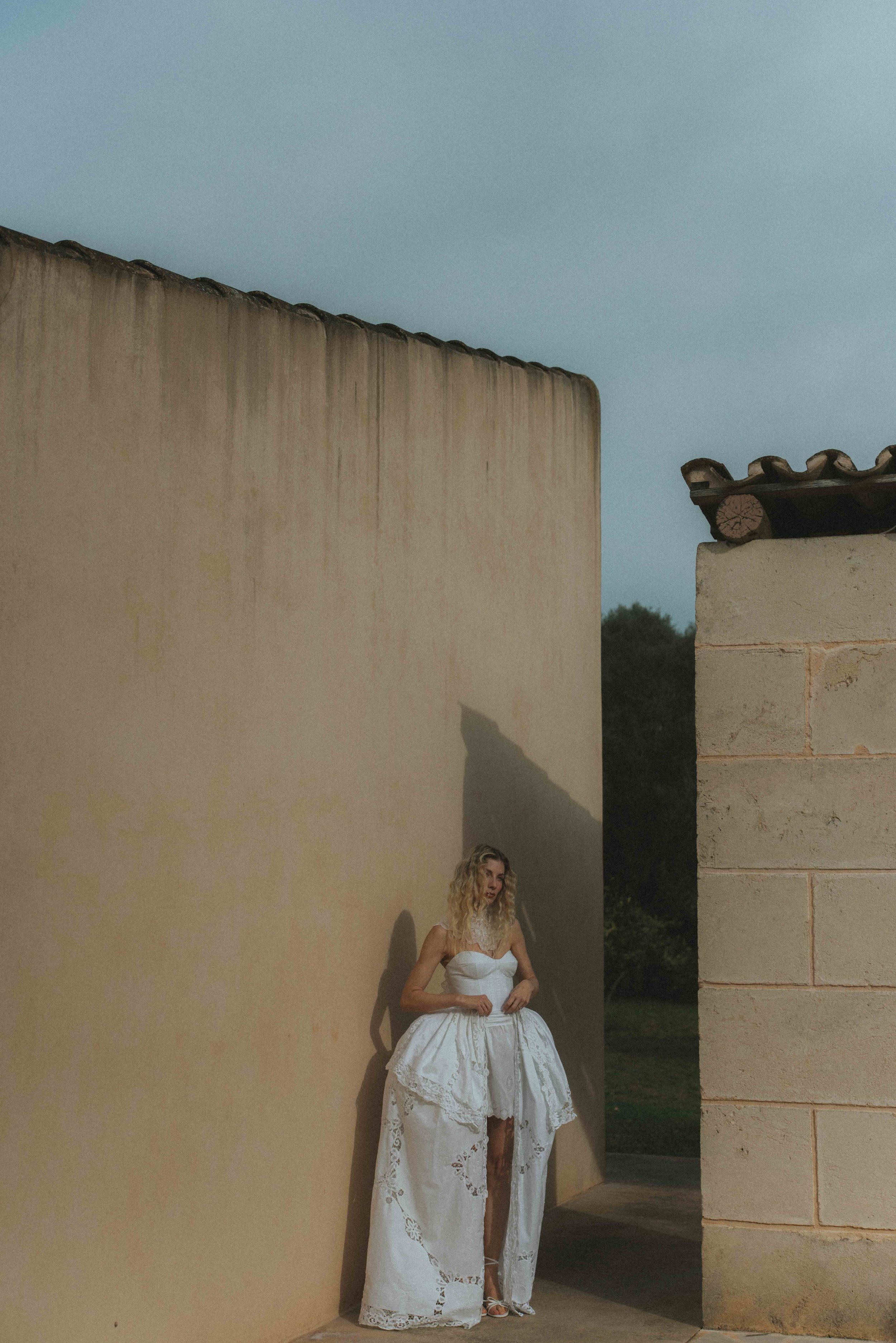 Frau in weißem Brautkleid steht vor einer hohen Mauer bei Sonnenuntergang, im Hintergrund Bäume und blauer Himmel.