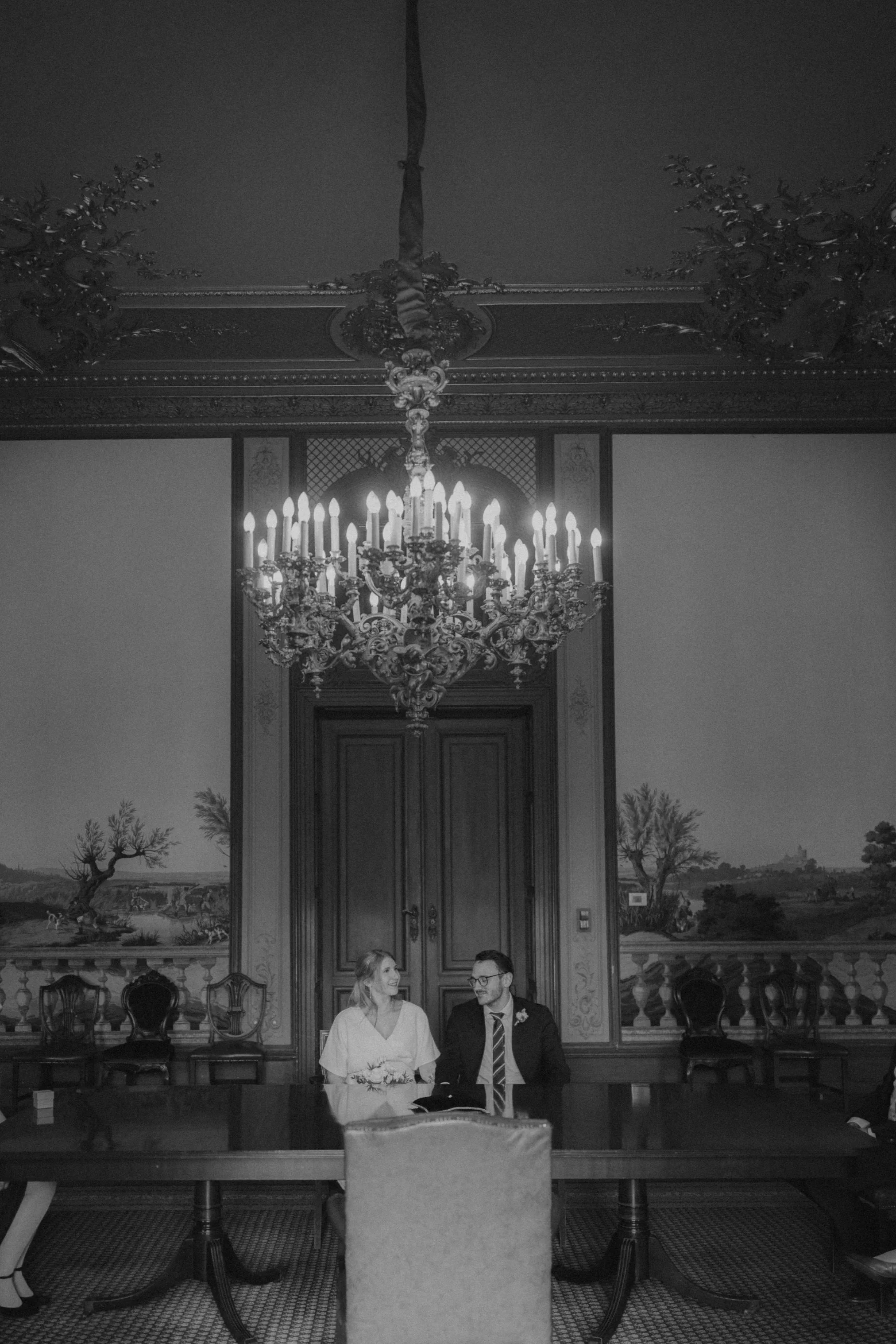 Ein elegantes Interieur mit einer großen Kristallleuchte, einem prunkvollen Kronleuchter, in einer luxuriösen, historischen Architektur, mit einem Brautpaar, das an einem langen Tisch sitzt.