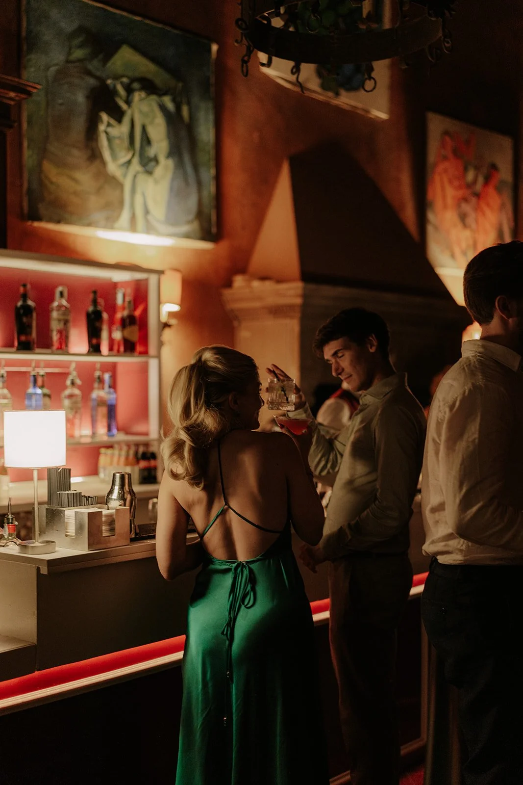 Menschen in einer Bar beim Anstoßen, im Hintergrund Kunstbilder und Flaschen an der Wand, gedämpftes Licht, elegantes Ambiente.