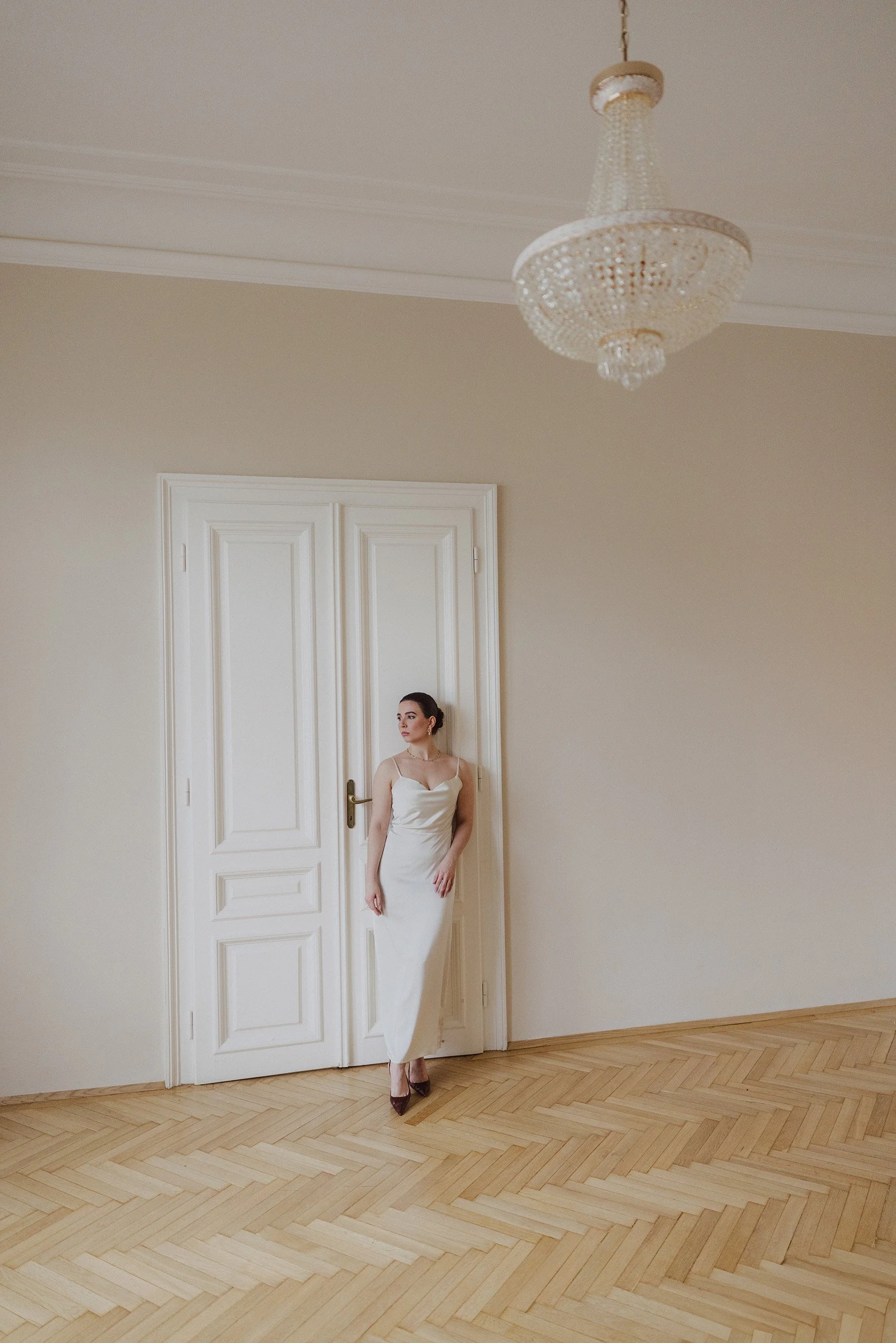 Eine Braut  in Braut Kleid steht in einem Raum mit hölzernem Boden und einer großen weißen Tür. Es hängt eine geschmückte Kronleuchter an der Decke. Wien. Altbau.