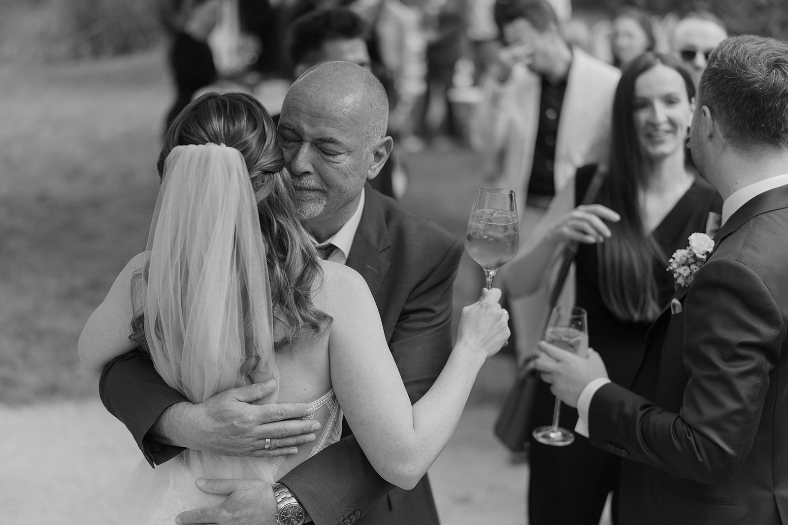 Ein Mann und eine Frau, vermutlich Braut und Vater, umarmen sich bei einer Hochzeit, während andere Gäste im Hintergrund bei einer Feier, bei der Sekt getrunken wird, lächelnd zusehen.