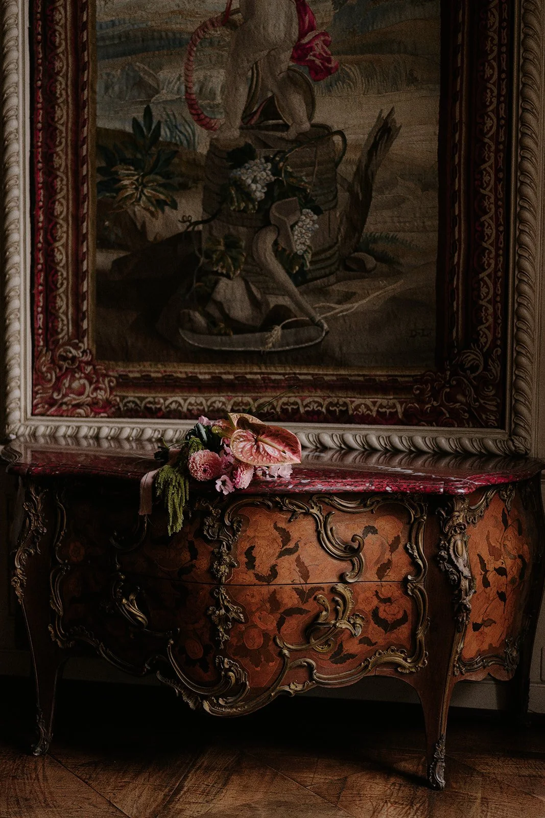 Ein antiker, kunstvoll verzierter Holztisch der St Charles Hall in Luzern mit floraler Mustertapete und einem Gemälde im Hintergrund. Auf dem Tisch liegt ein Strauß aus bunten Blumen.