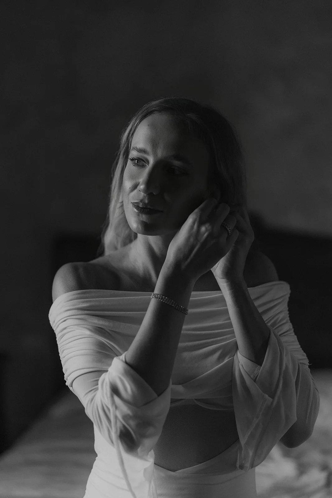 Schwarze und weiße Nahaufnahme einer Frau beim Anpassen eines Ohrrings, mit off-shoulder Kleid, entspannte Pose, sanftes Licht, nachdenklicher Ausdruck.