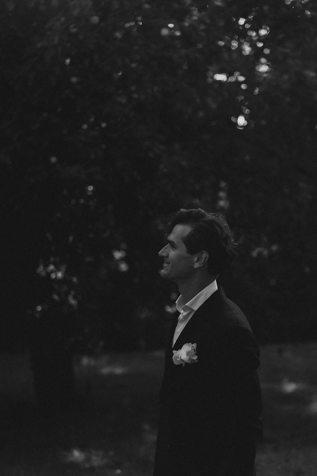 Schwarz-weiß-Foto eines Bräutigams in einem Anzug mit Blume im Revers, der im Freien bei Dämmerung nach rechts lächelt, mit Bäumen im Hintergrund.