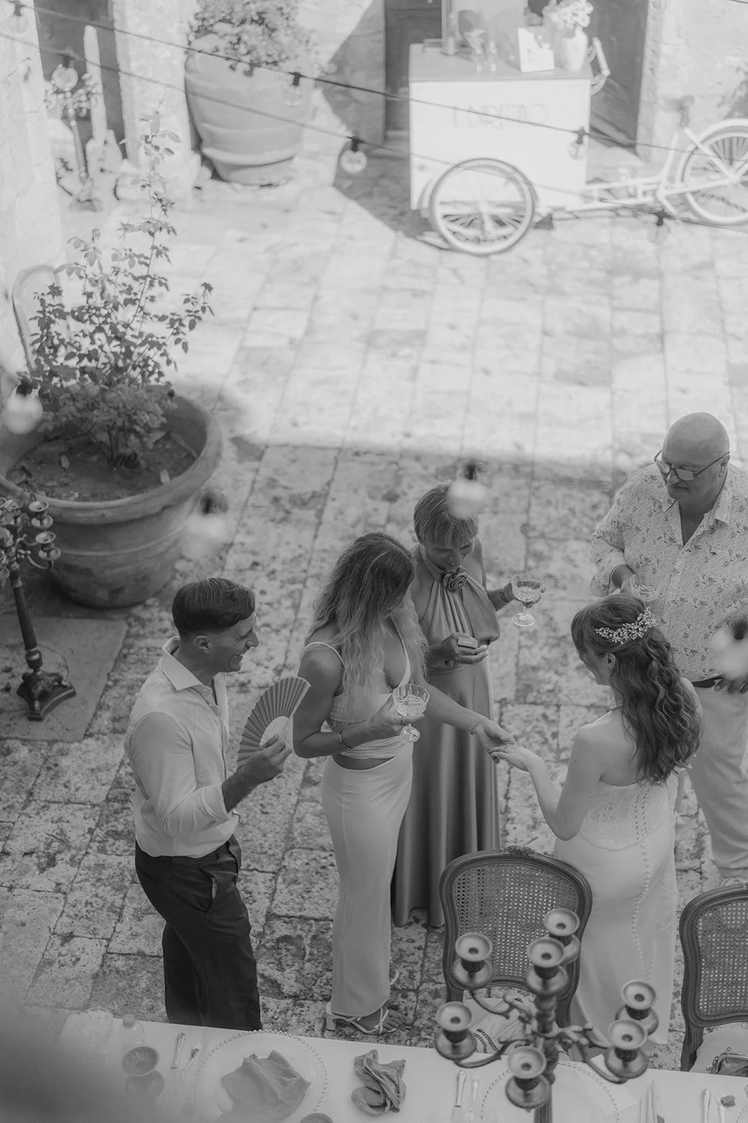 Menschen bei einer Hochzeit im Innenhof, eine Frau in weißem Kleid wird mit einer Hand berührt. Es gibt eine Gruppe von fünf Personen, zwei Frauen und drei Männer, die Getränke halten, während sie miteinander sprechen. Die Szene ist festlich dekorier